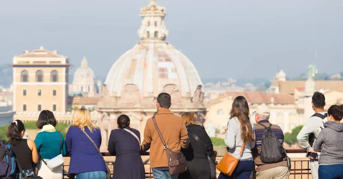 A group of tourists at the ledge of an overlook as they stare out at Rome, Italy from a distance. It's sunny outside.