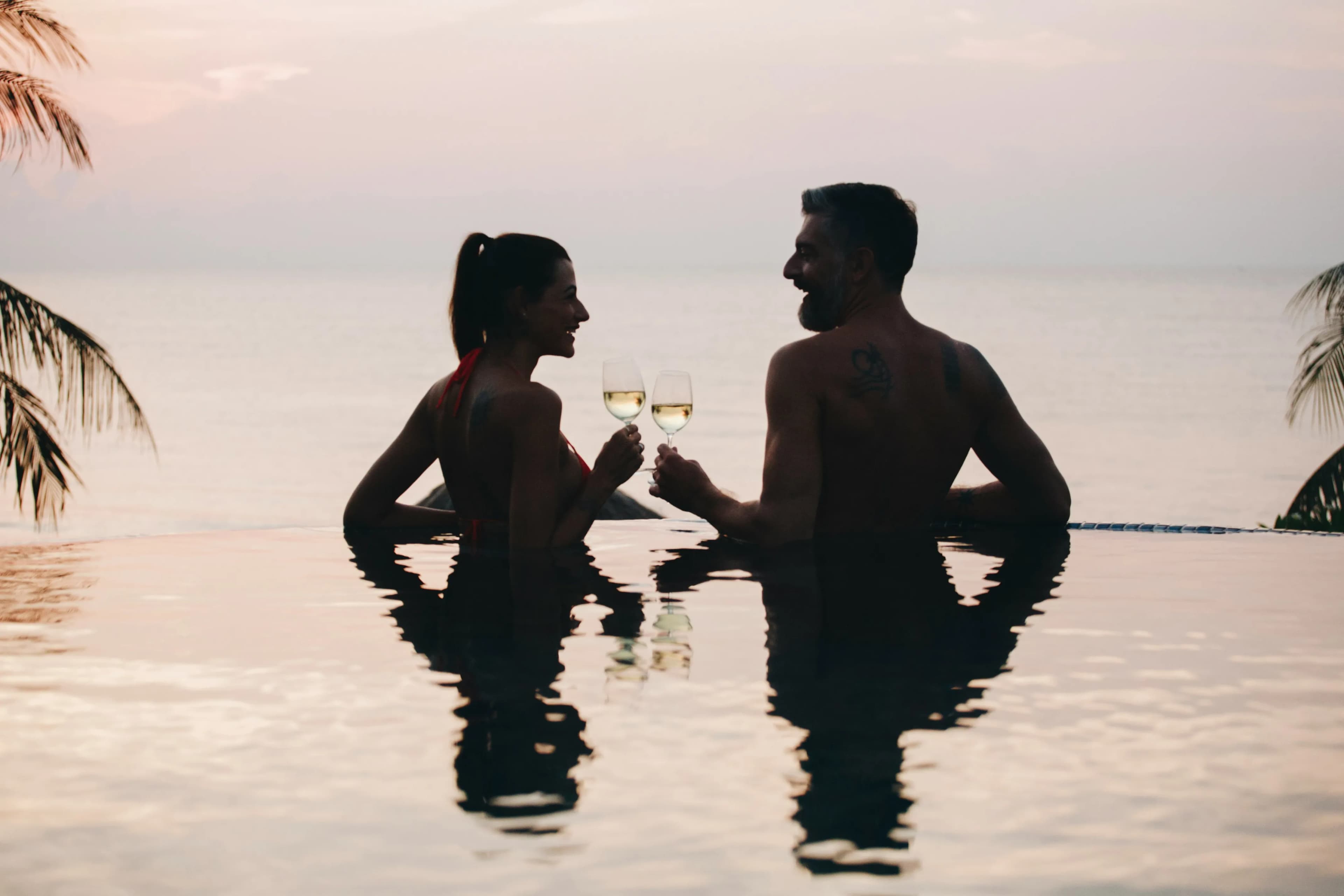 A couple in swimwear enjoys glasses of white wine while relaxing in an infinity pool at sunset, with the ocean and palm trees in the background—capturing the essence of Top Honeymoon Destinations 2024.
