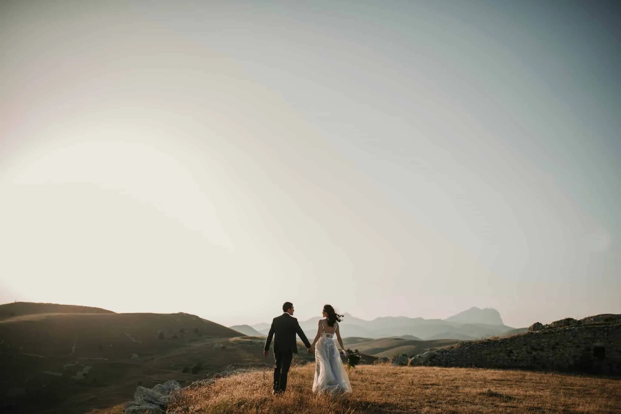 A couple in formal attire walks hand in hand on a grassy hillside at sunset, reminiscent of a private group tour admiring nature’s beauty. The expansive landscape of rolling hills and distant mountain range under a clear sky creates a romantic and serene atmosphere.