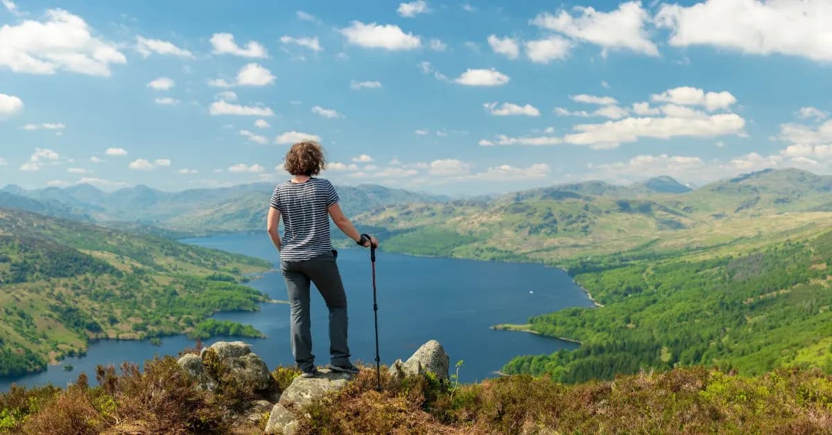 A hiker with trekking poles stands on a rocky heather-covered summit looking over a long blue loch winding between green Scottish hills under a bright sky.
