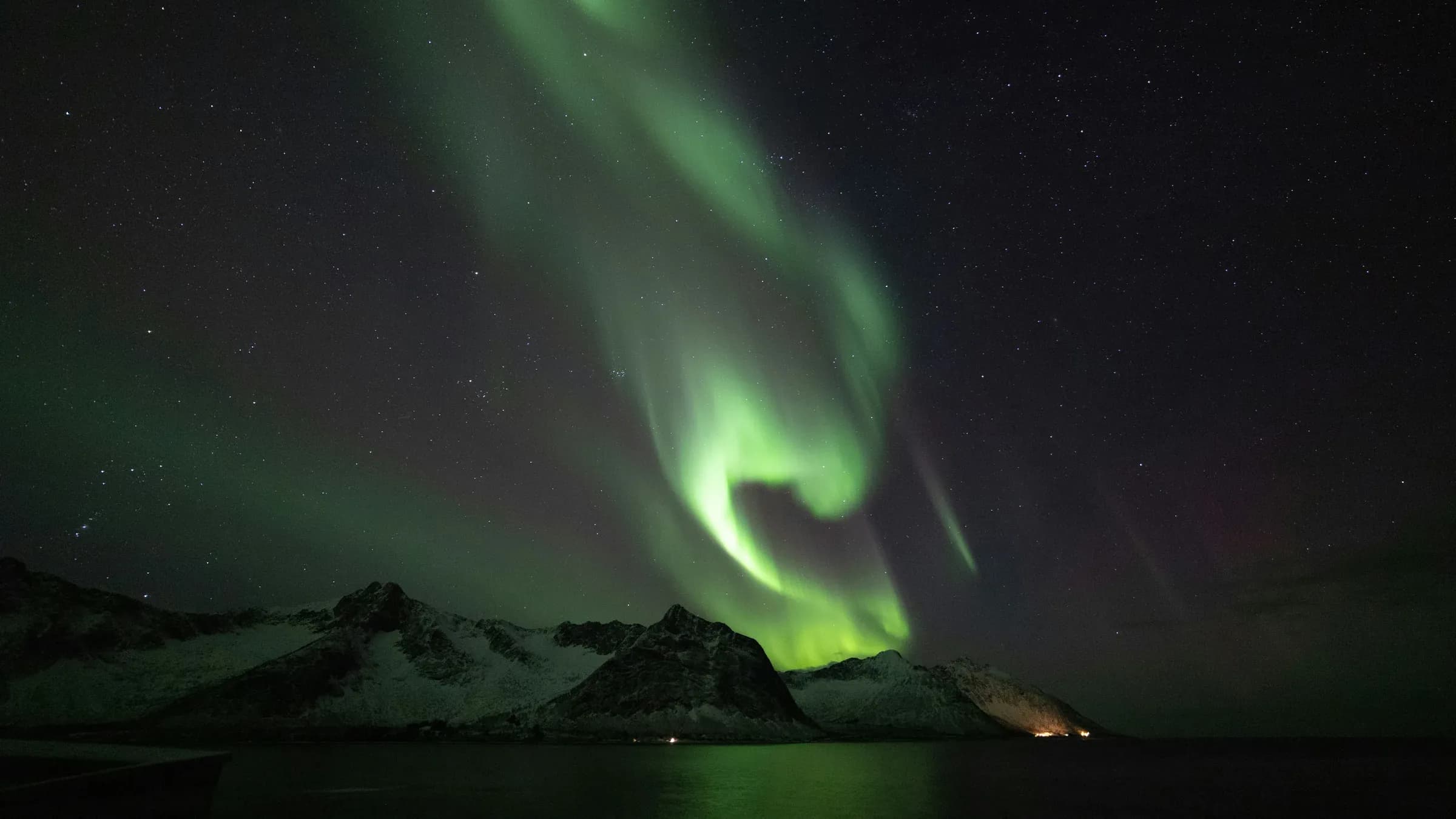 Green aurora curling across a star-filled night sky above snow-covered coastal mountains in northern Norway, its light reflected on still dark water.