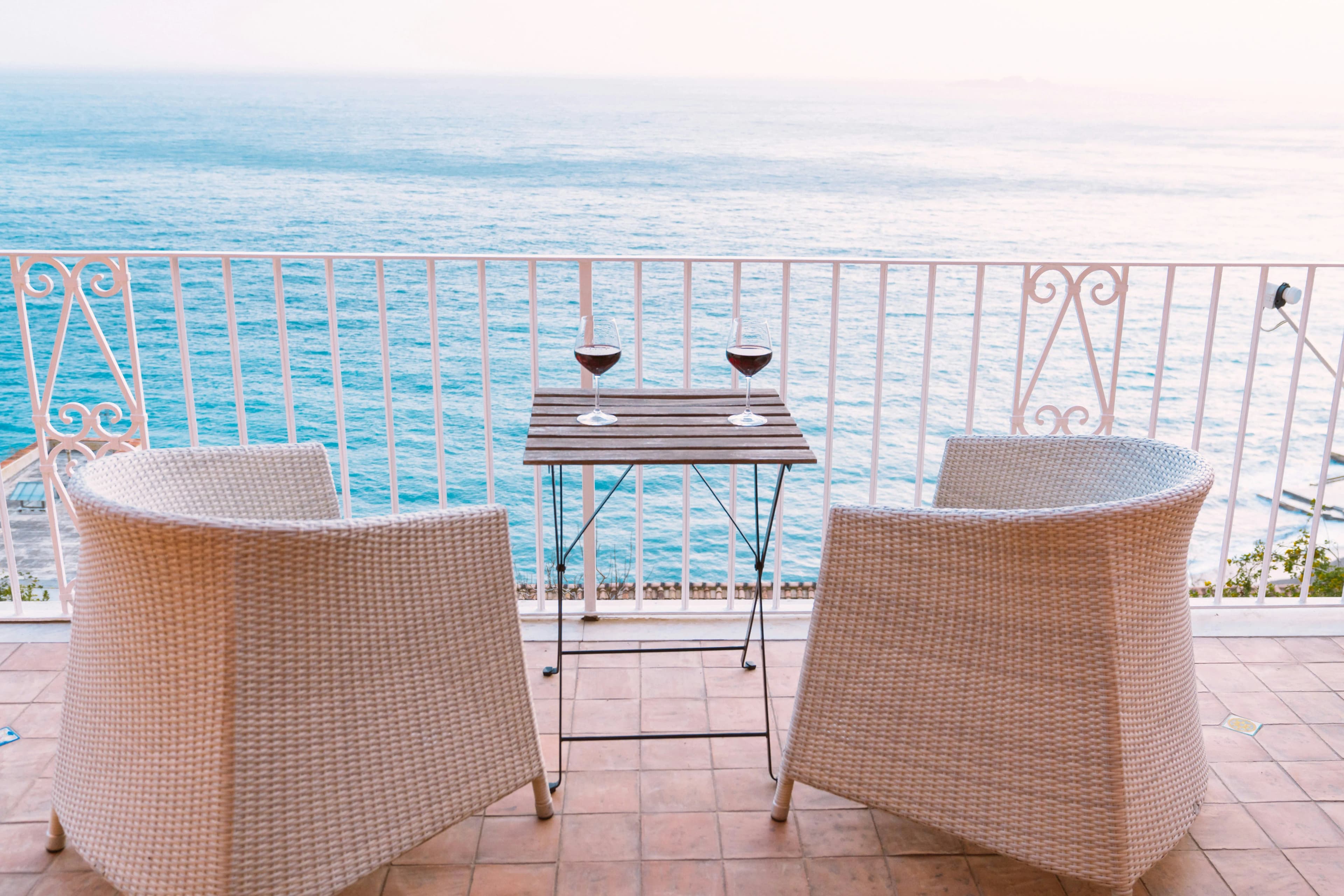 Two wicker chairs face a small wooden table with two glasses of red wine on a tiled balcony, overlooking the calm, blue ocean under a pale sky—perfect for reflecting on what is Victory Day in Croatia and its meaning.