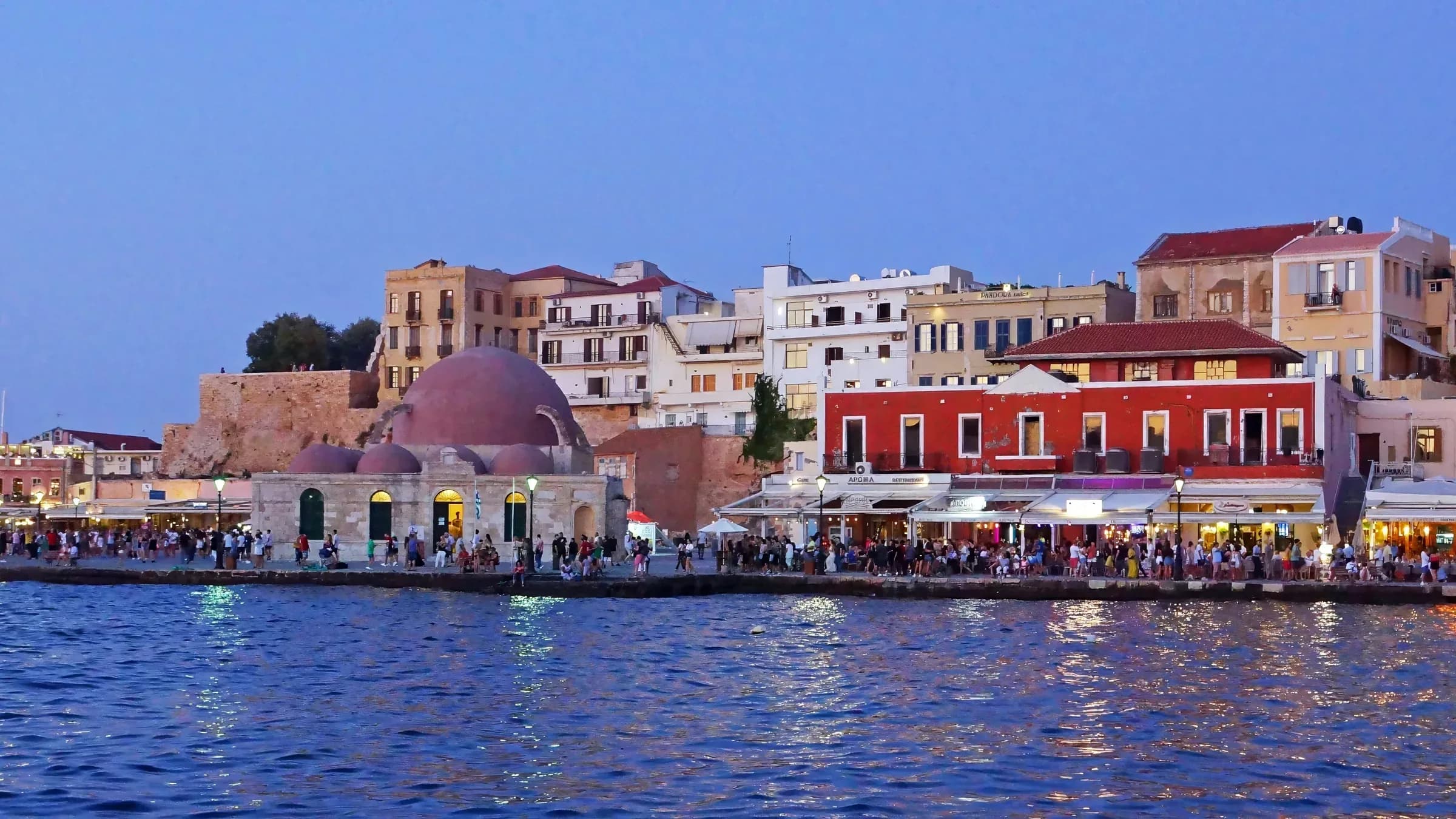 The Venetian harbour at Chania, Crete at dusk, the domed Kucuk Hasan mosque and lamplit waterfront tavernas reflected in calm water with people along the quay.