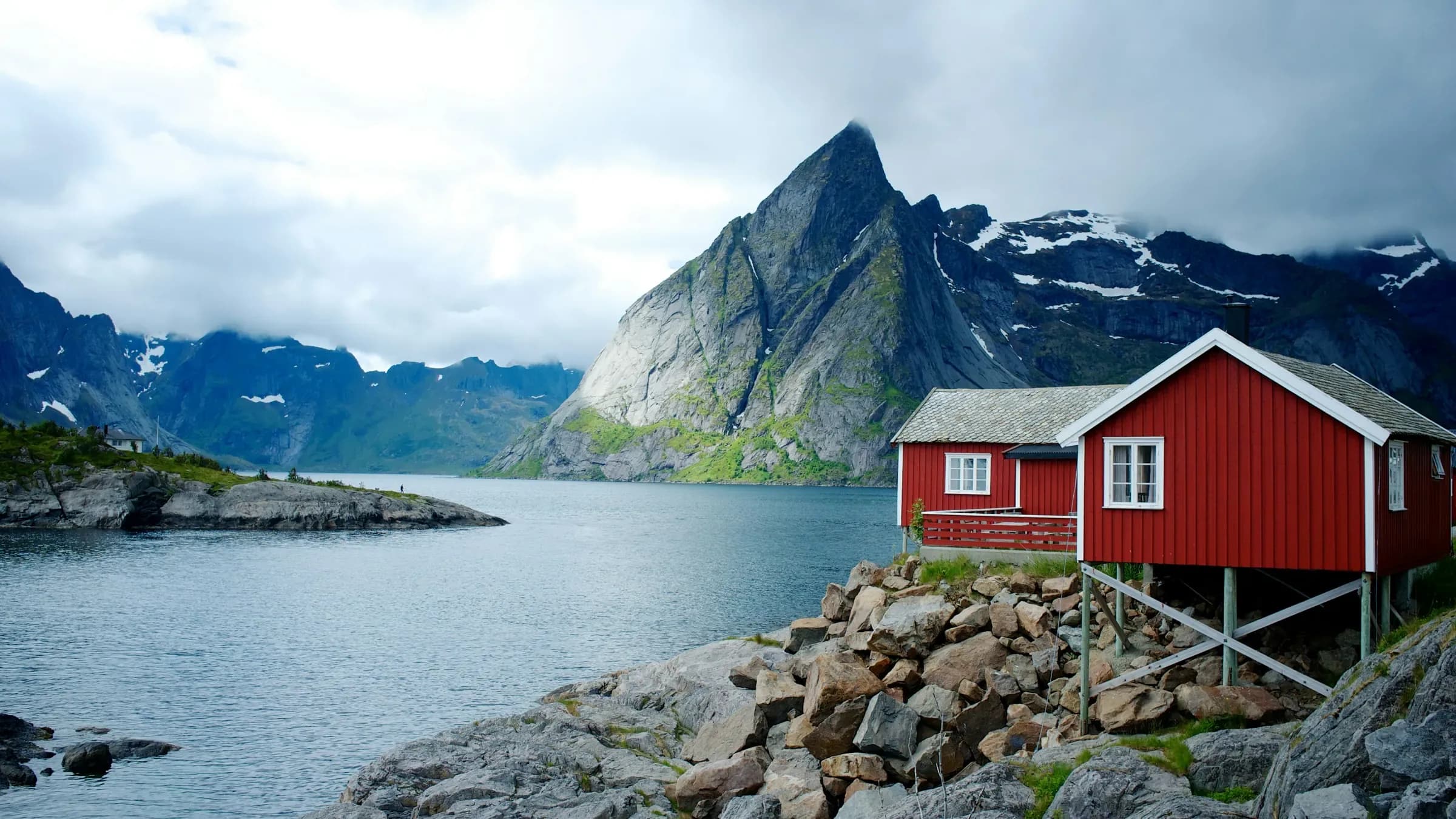 A red timber rorbu cabin on stilts at the edge of a Lofoten fjord, a sharp granite peak and snow-streaked mountains rising across the water under heavy cloud.