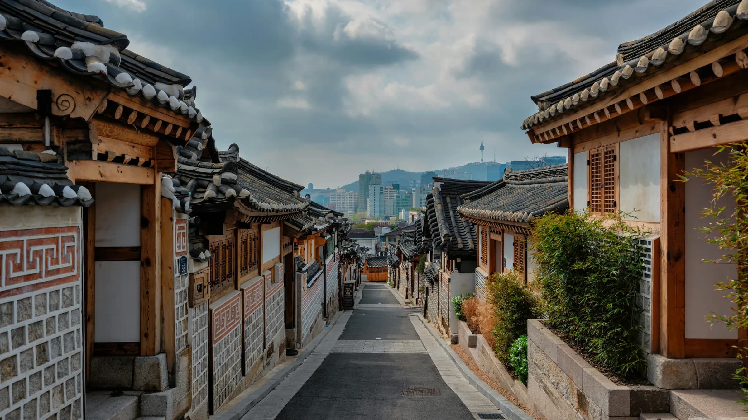 A narrow lane between traditional tiled-roof hanok houses in Seoul, with city hills beyond.