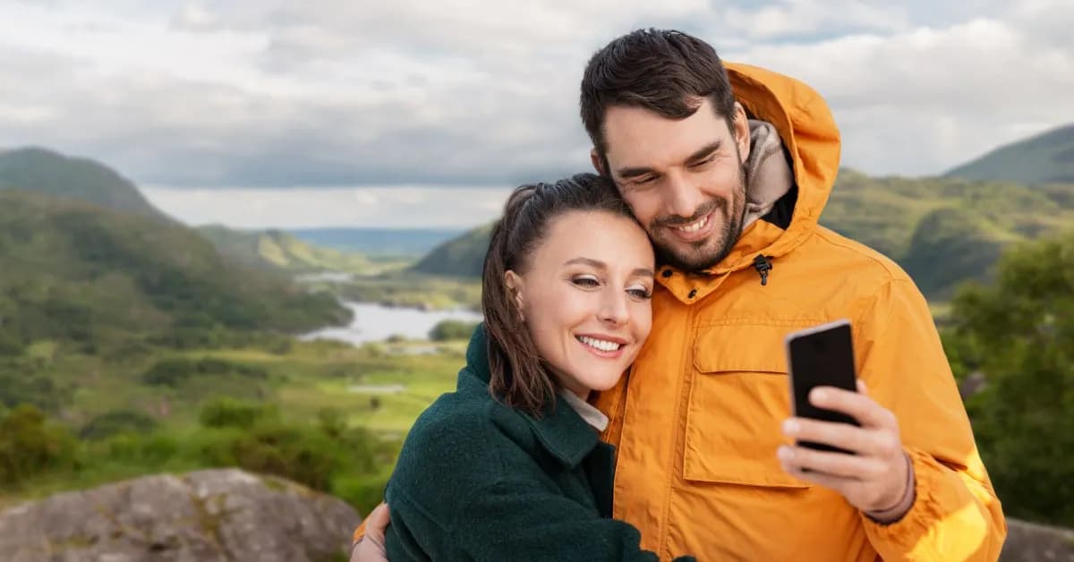 A smiling couple in rain jackets taking a selfie above the lakes and mountains of Killarney National Park