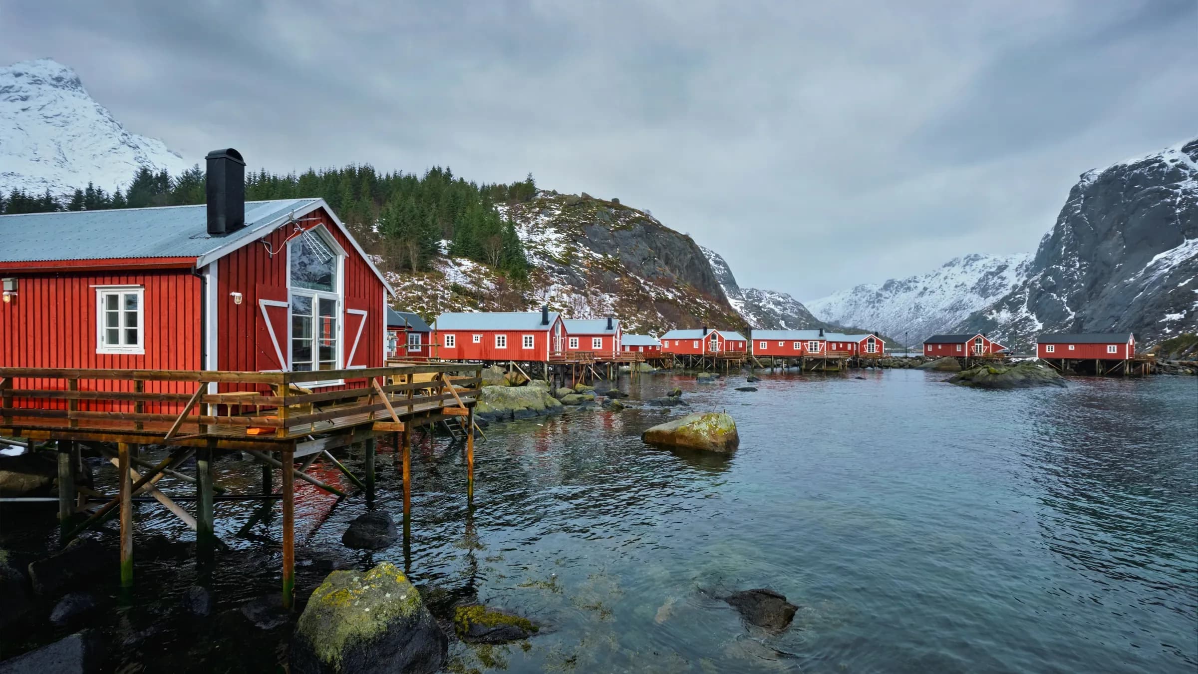 A row of red rorbuer on wooden stilts curving along a sheltered Lofoten inlet, snow on the surrounding hills and clear shallow water in the foreground.