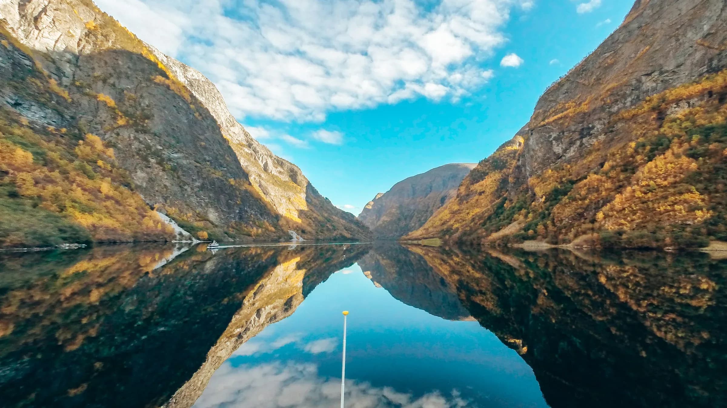 The view forward from a small boat down the Nærøyfjord in autumn, steep golden-birch walls on either side mirrored in glassy water with a single vessel ahead.