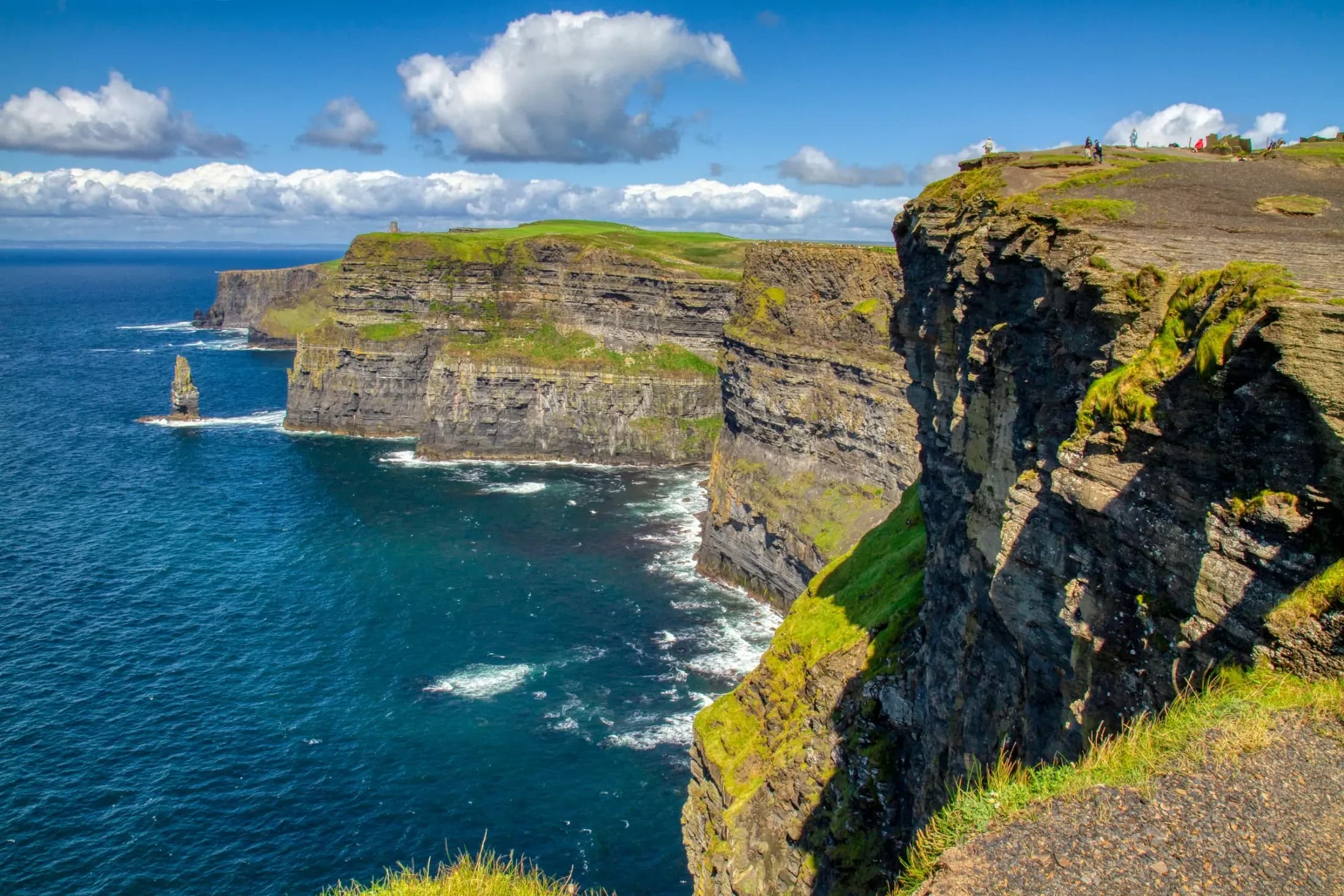 The Cliffs of Moher in County Clare rising in layered green-topped sea cliffs above the deep blue Atlantic, with a lone sea stack offshore under a bright sky.
