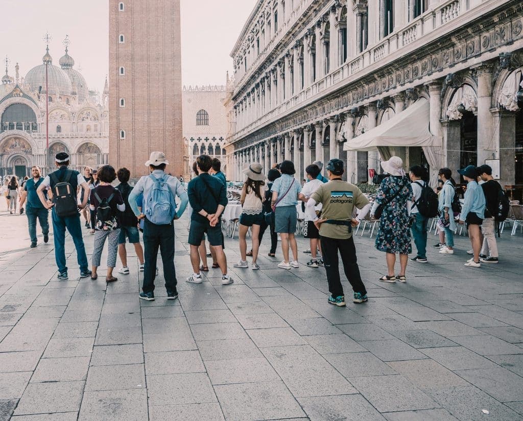 A group of tourists enjoys the benefits of escorted tours in a historic square, surrounded by ornate buildings. In the background is a tall brick tower, likely part of a cathedral. The scene is bright, suggesting daytime, and people wear casual, summer clothing.