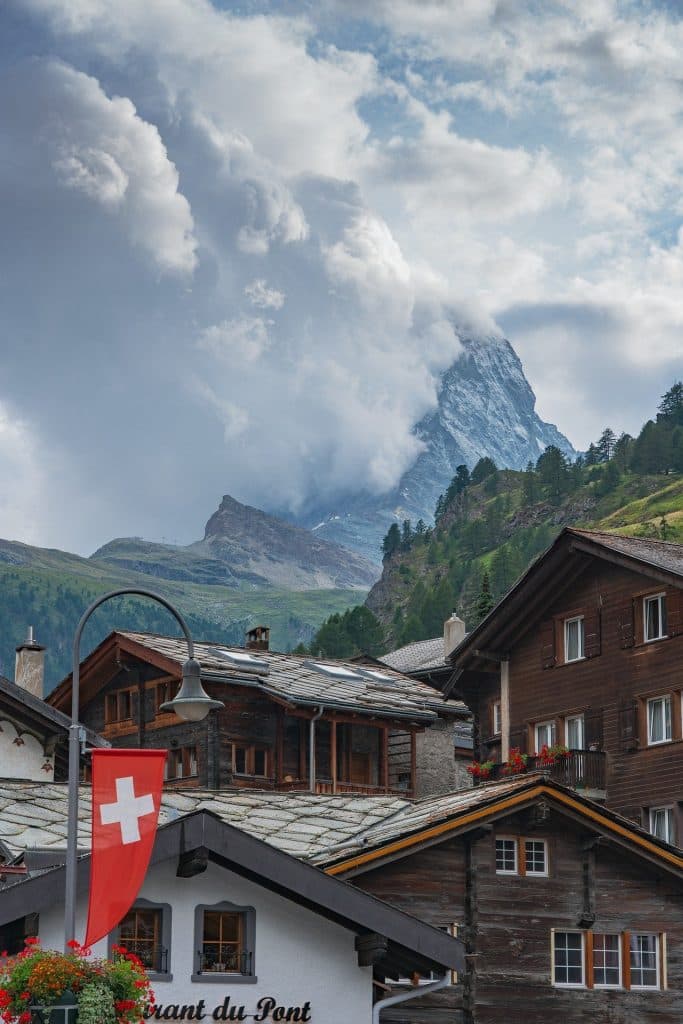 Swiss village scene with traditional wooden houses, one displaying a Swiss flag. The background features a dramatic view of the Matterhorn partially obscured by clouds. Red flowers decorate the balconies, adding color to the rustic setting.