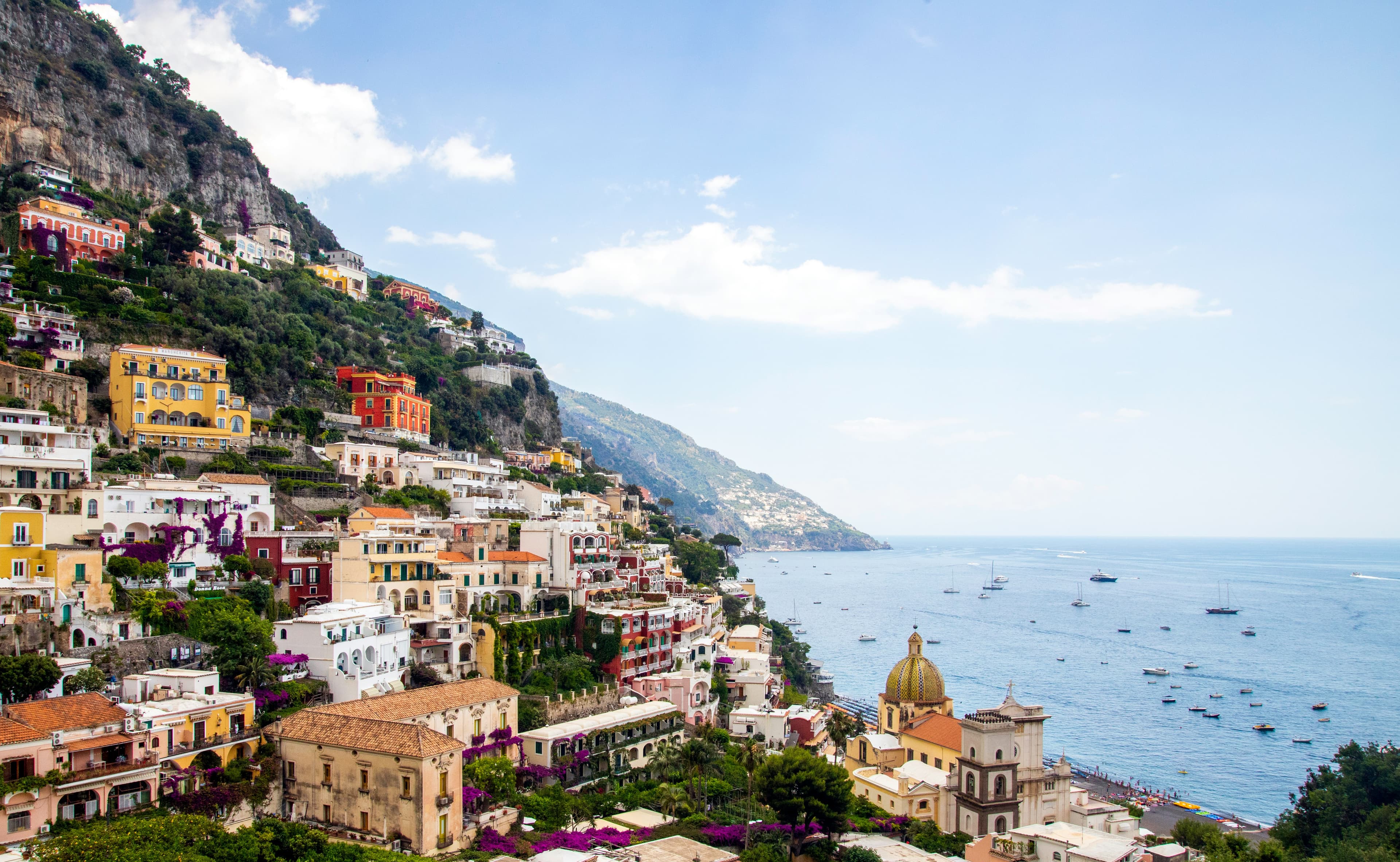 Colorful hillside buildings overlook the blue sea in Positano, Italy, with boats scattered in the water—echoing the dramatic cliffs found near Greece's Mount Olympus—lush greenery thrives under a bright, partly cloudy sky.