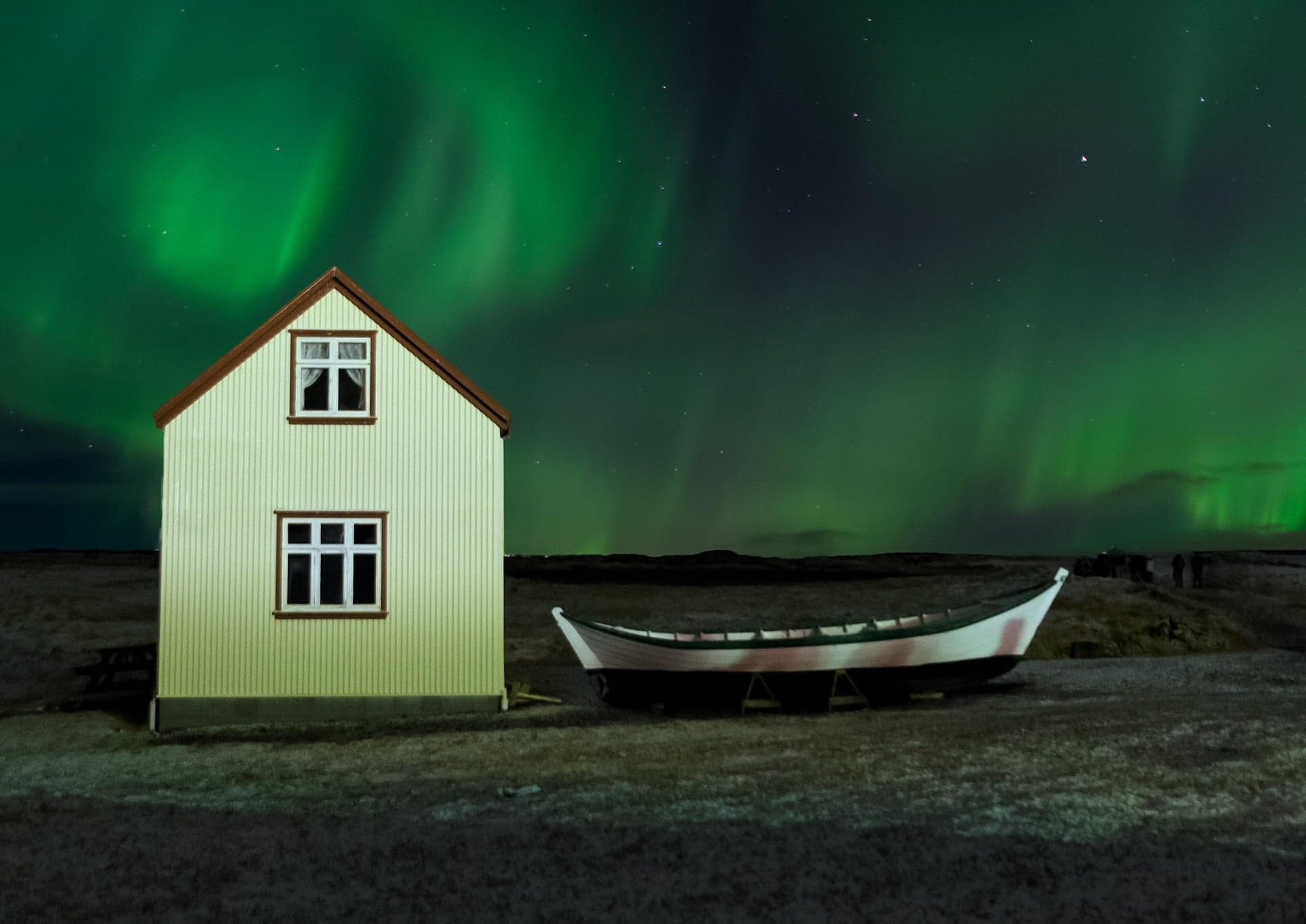 A small yellow house and a white boat sit on a grassy field under a night sky illuminated by vibrant green northern lights, creating a scene that might spark questions like, "Does Greece celebrate Halloween?.