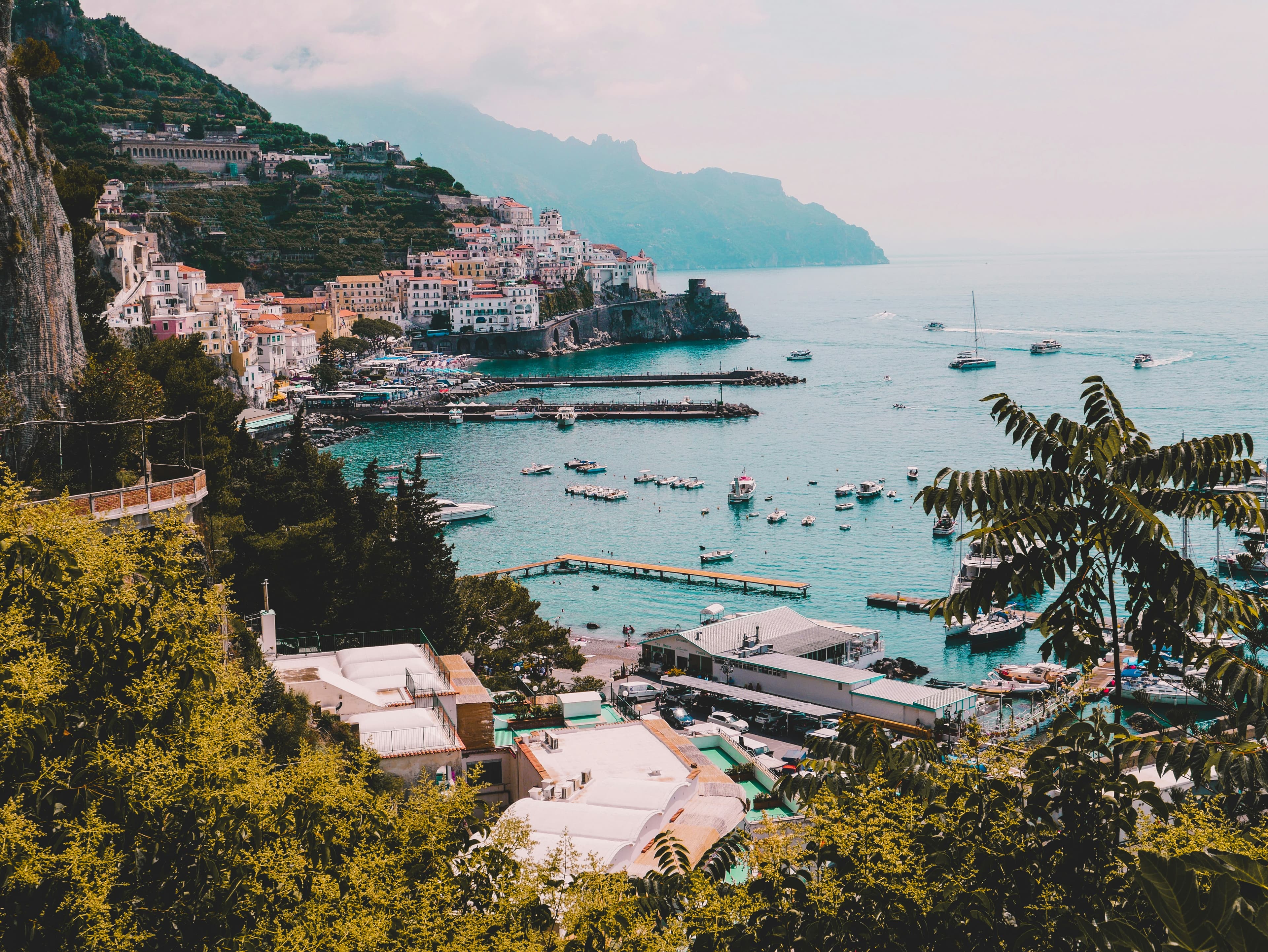 Coastal town with colorful buildings on steep hills, overlooking a turquoise harbor dotted with boats and yachts; lush greenery in the foreground and mountains in the hazy distance—perfect for a romantic Spain and Portugal Honeymoon escape.