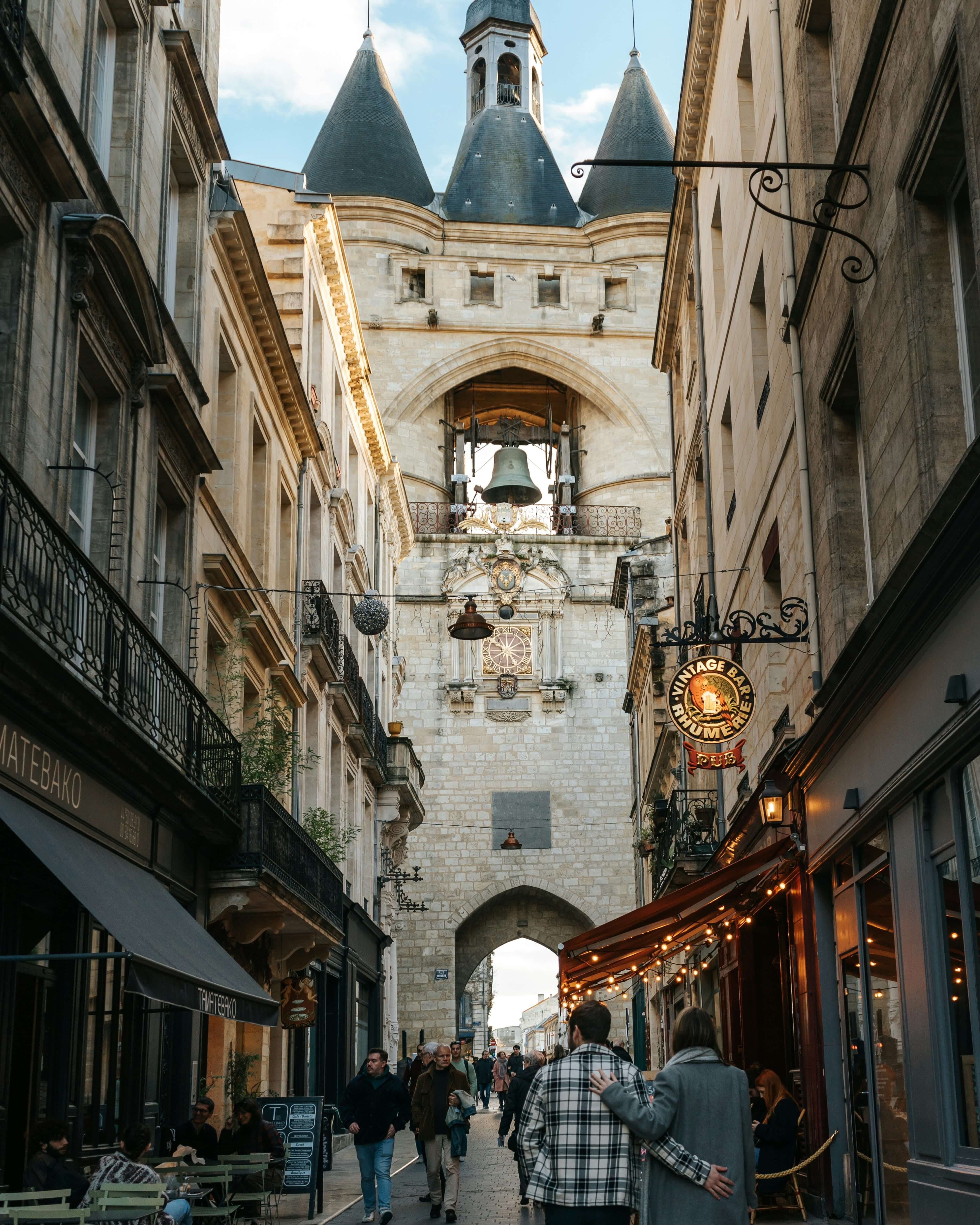 A stone street with outdoor cafes leads to a large historic clock tower with a bell and spires—an idyllic setting for an Ireland or Scotland honeymoon. People walk beneath the archway, as warm lights glow from the shops and restaurants.