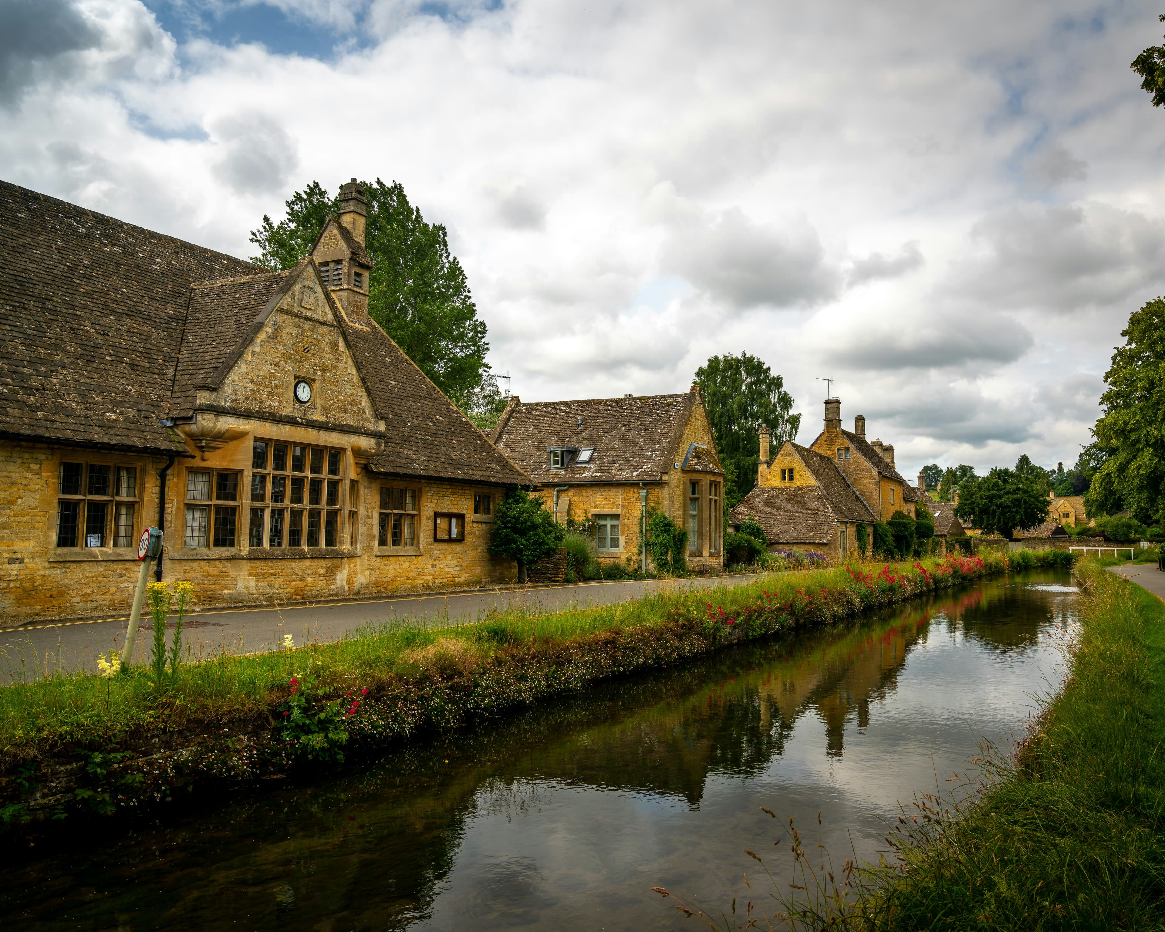 A tranquil village scene with stone buildings, a narrow river reflecting the cloudy sky, and green trees lining the water’s edge in the countryside.