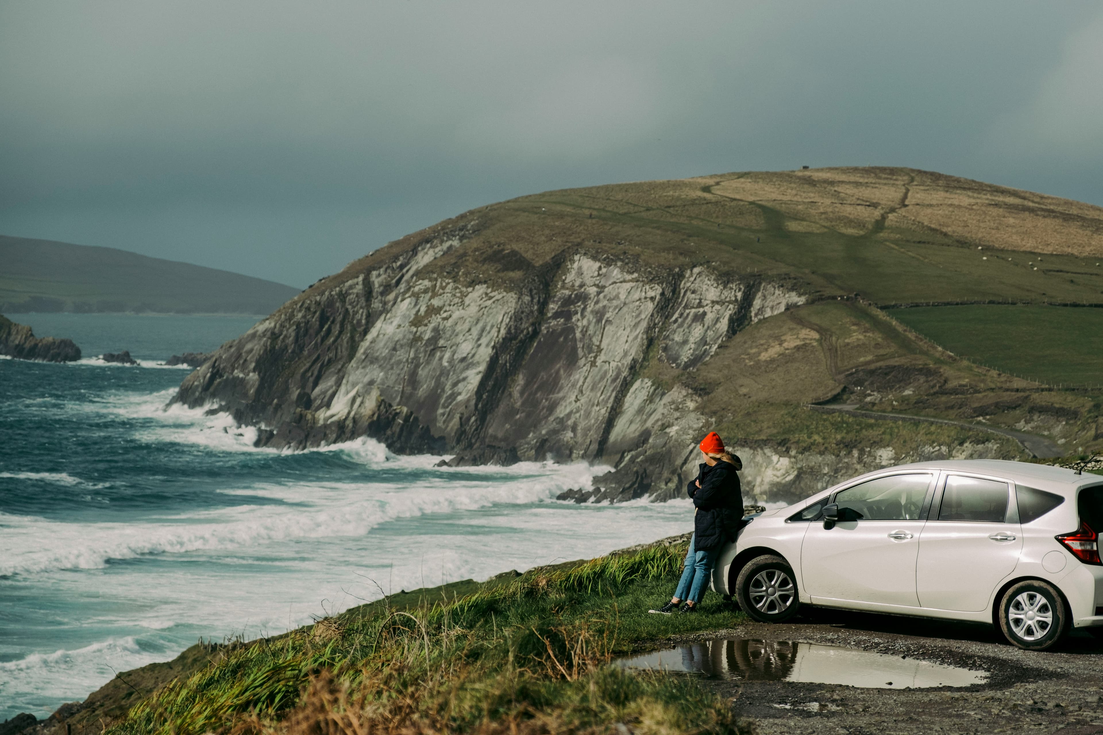 A person in a red hat stands beside a white car, gazing at waves crashing against rocky cliffs under a cloudy sky on a grassy coastal road—an ideal setting for Azores Honeymoon Packages.