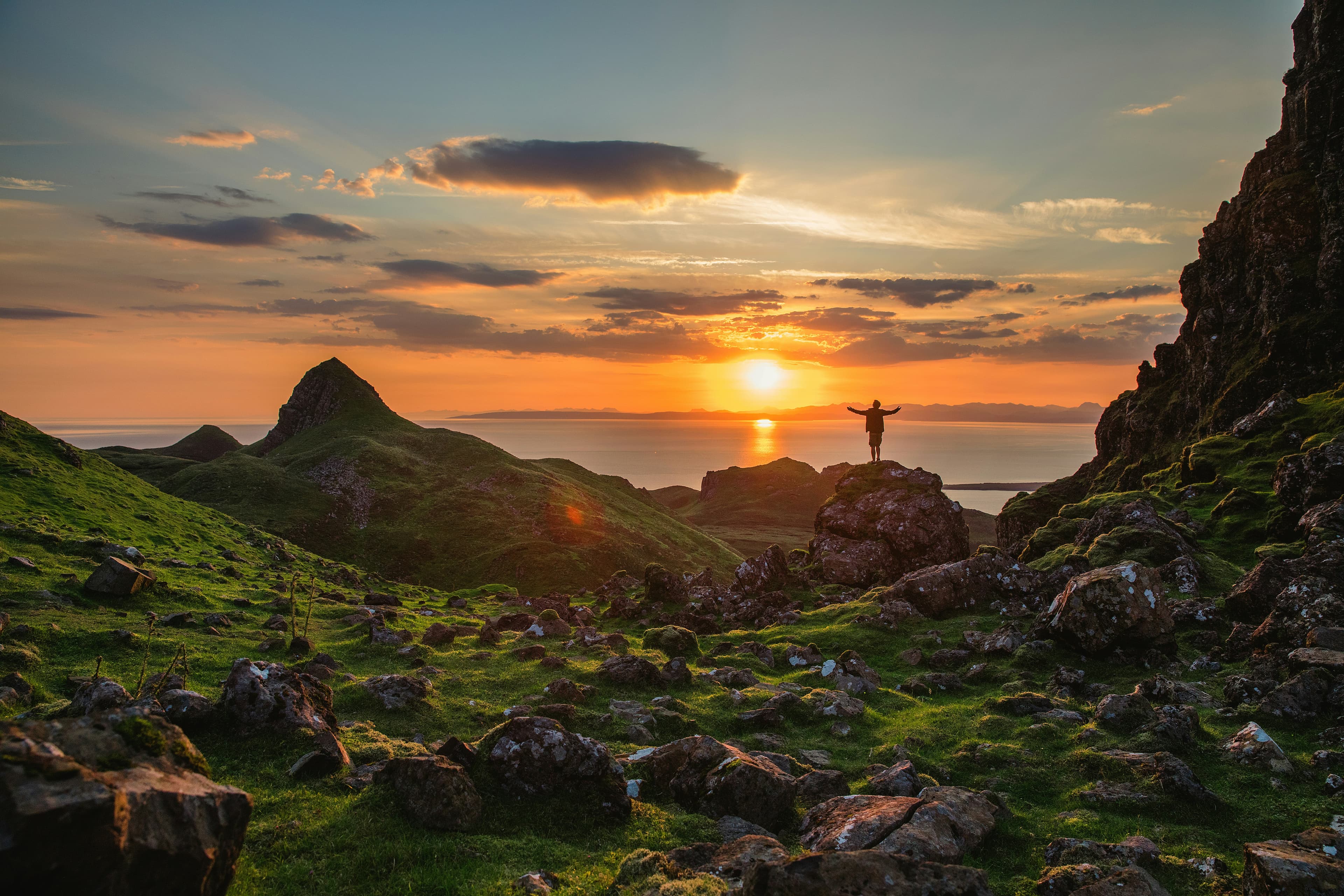 A person stands on a rocky hill with arms raised, surrounded by green hills and rocks, watching a colorful sunset over the sea and distant mountains.