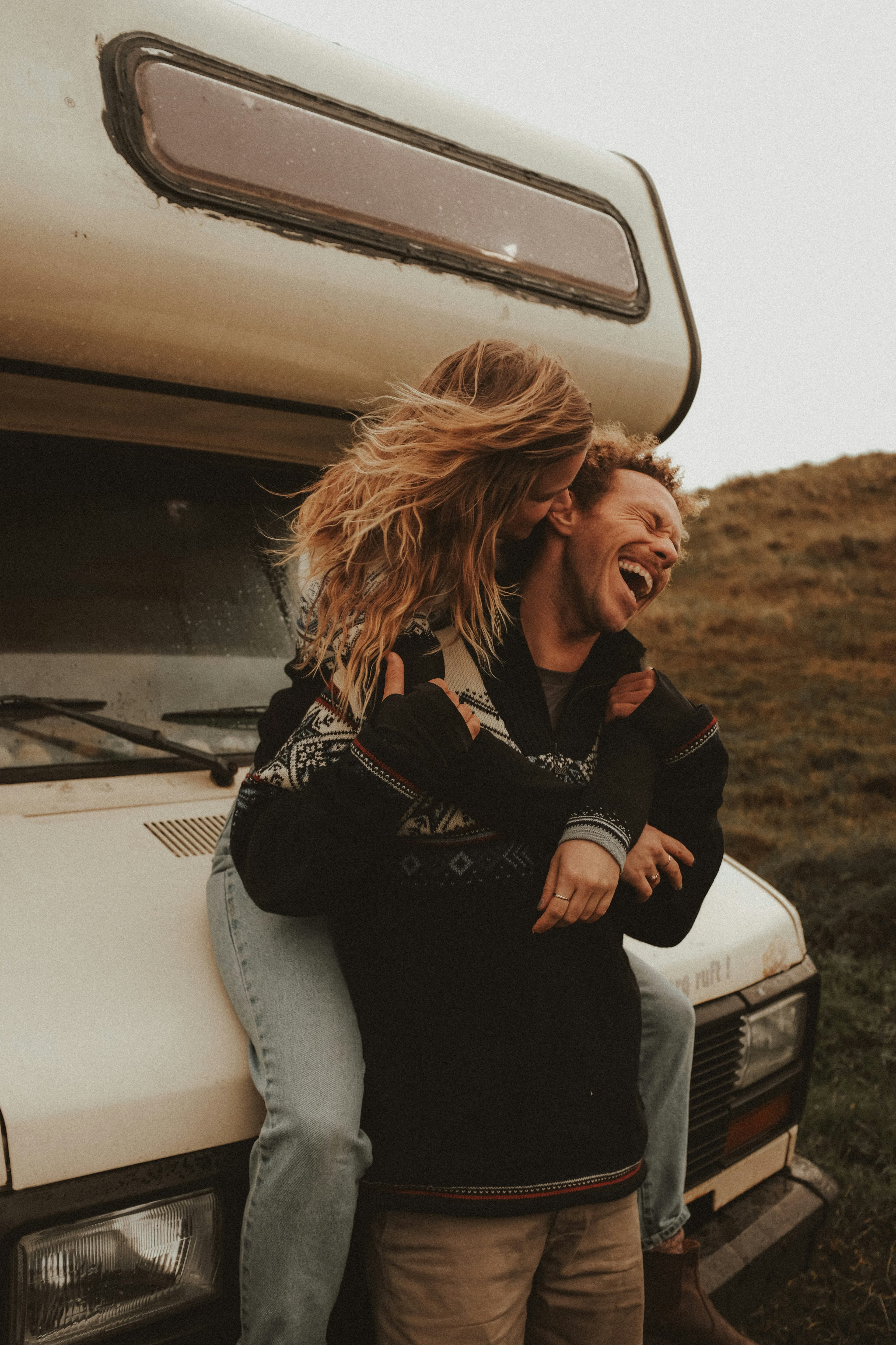 A woman with long hair playfully hugs a laughing man from behind as they sit on the front of a camper van parked in a grassy outdoor setting, dreaming about future adventures and Ryder Cup 2027 travel packages. Both appear happy and relaxed.