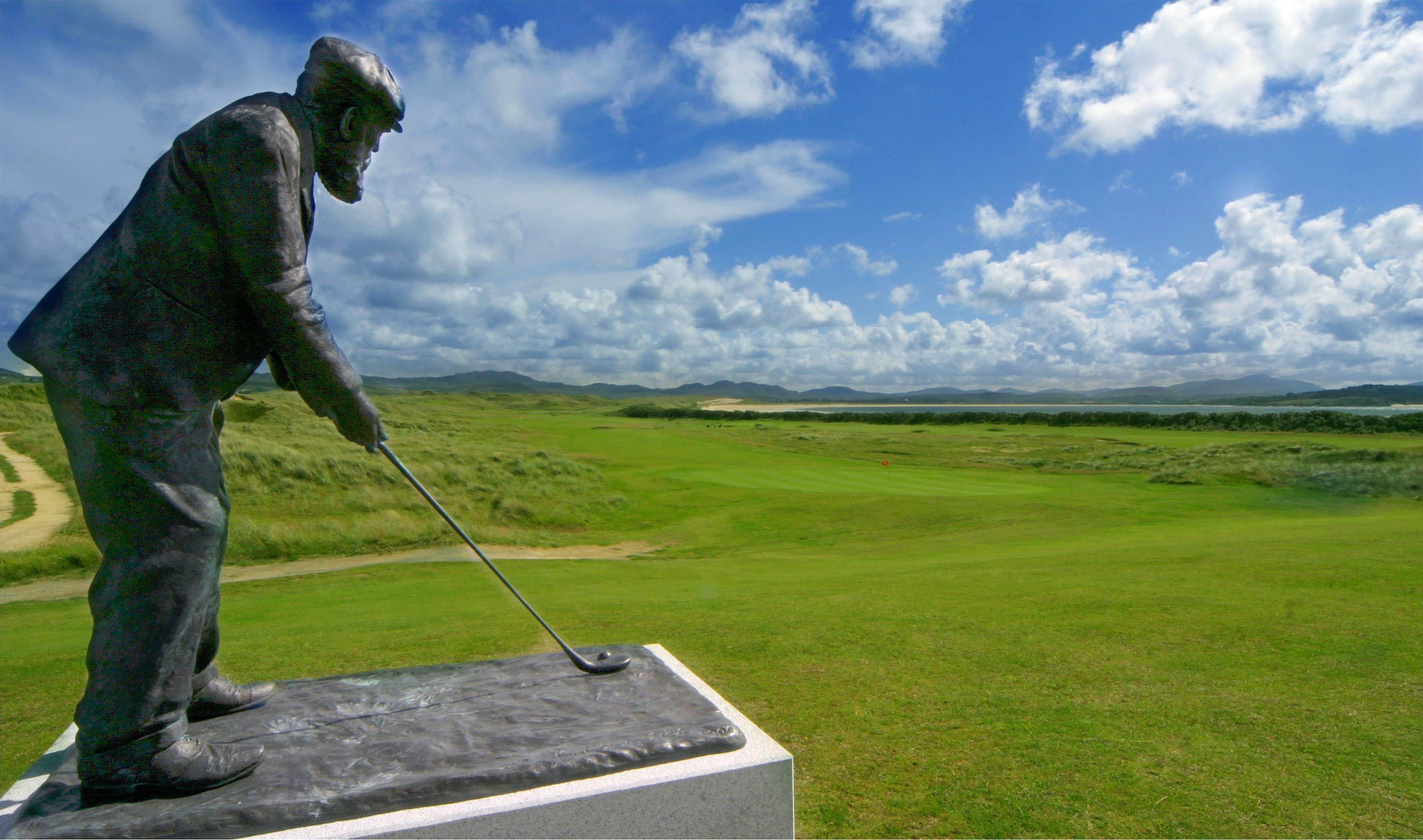 A bronze statue of a golfer preparing to putt stands on a pedestal overlooking a lush green golf course, reminiscent of the landscapes visited by Steelers in Ireland, with rolling hills and a partly cloudy blue sky in the background.