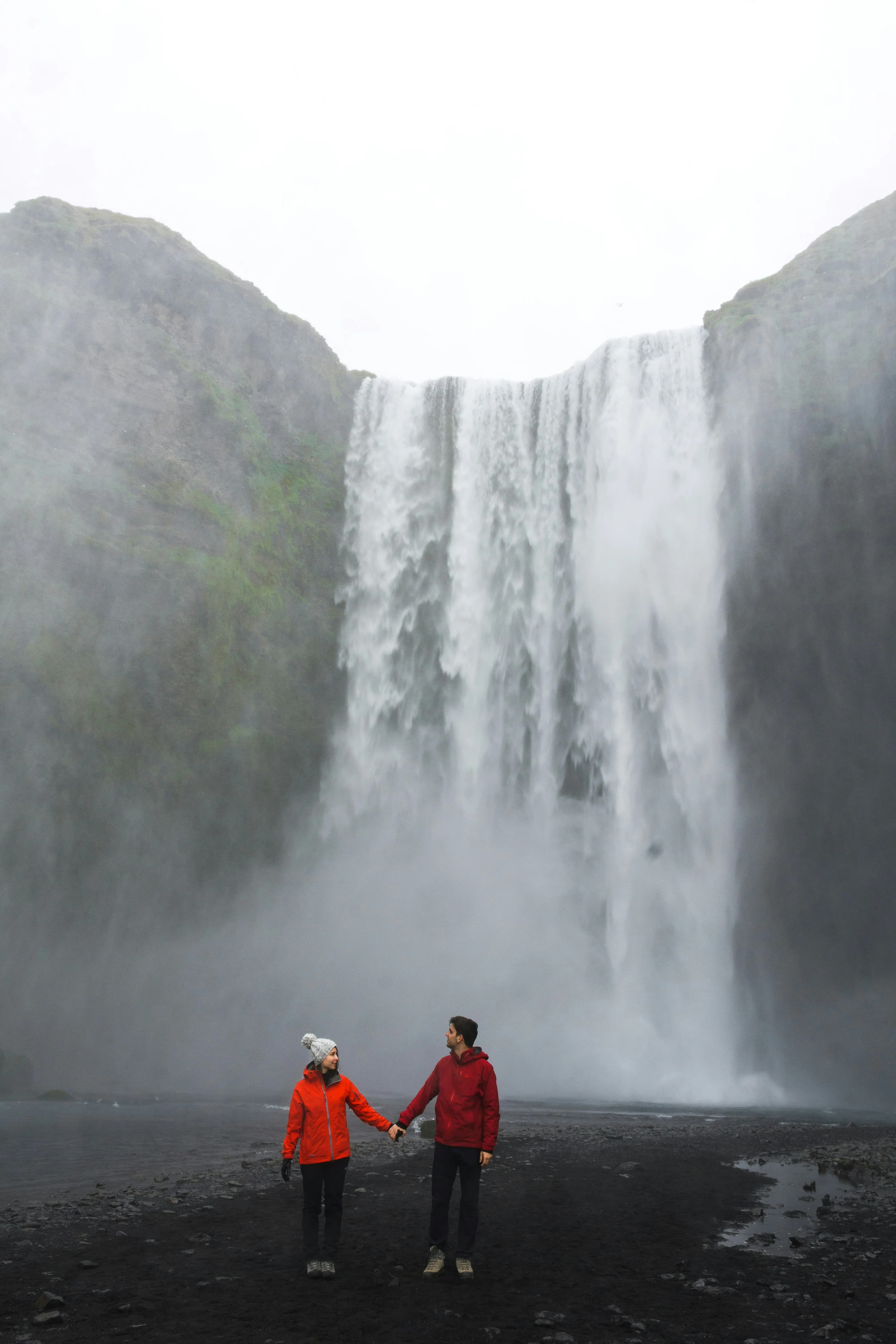 Two people in red jackets hold hands before a misty waterfall, rocky cliffs, and cloudy sky—an enchanting scene inspired by a Guide to Honeymoon in France.
