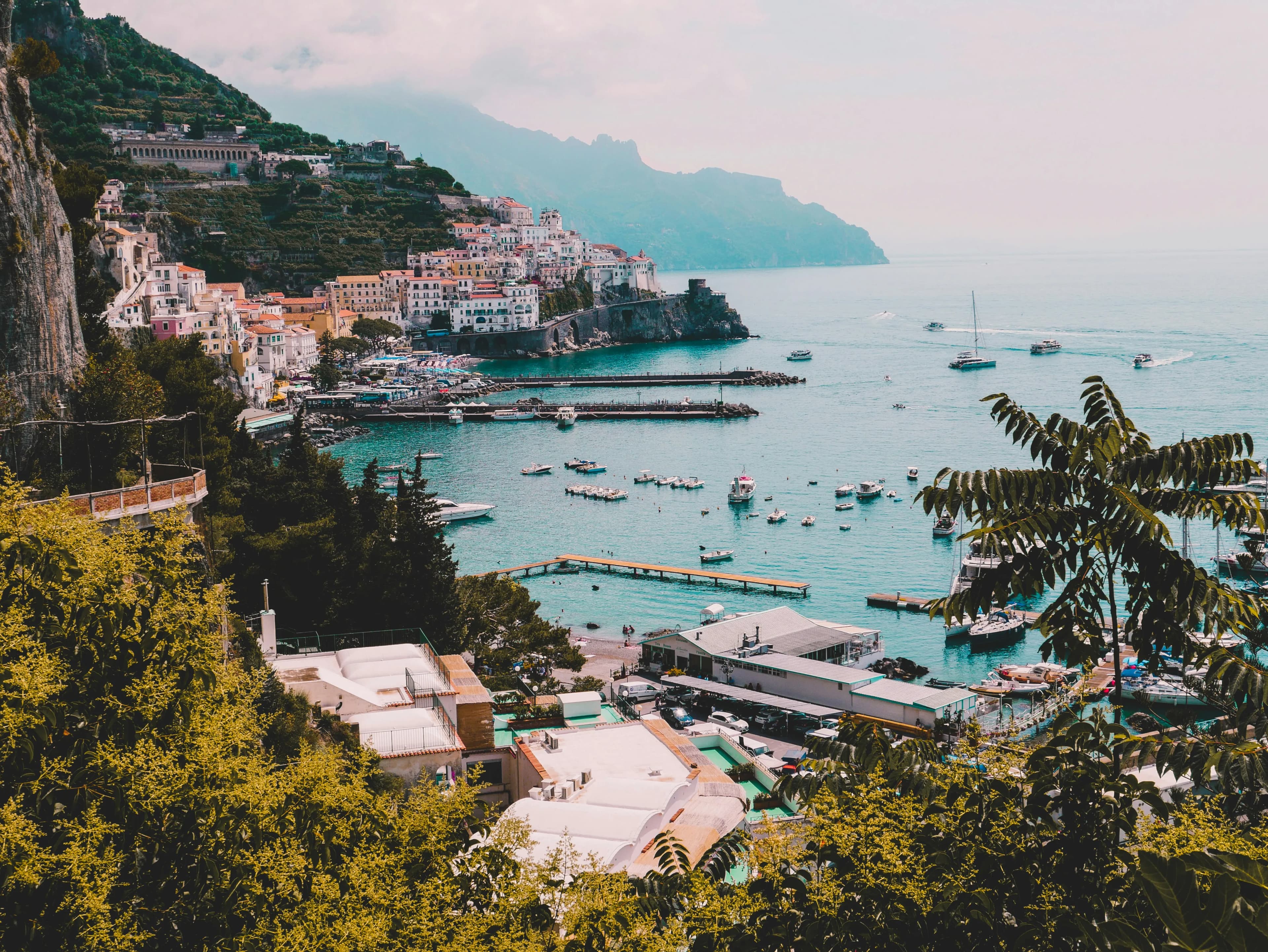 Coastal town with colorful buildings on steep hills, overlooking a turquoise harbor dotted with boats and yachts; lush greenery in the foreground and mountains in the hazy distance—perfect for a romantic Spain and Portugal Honeymoon escape.