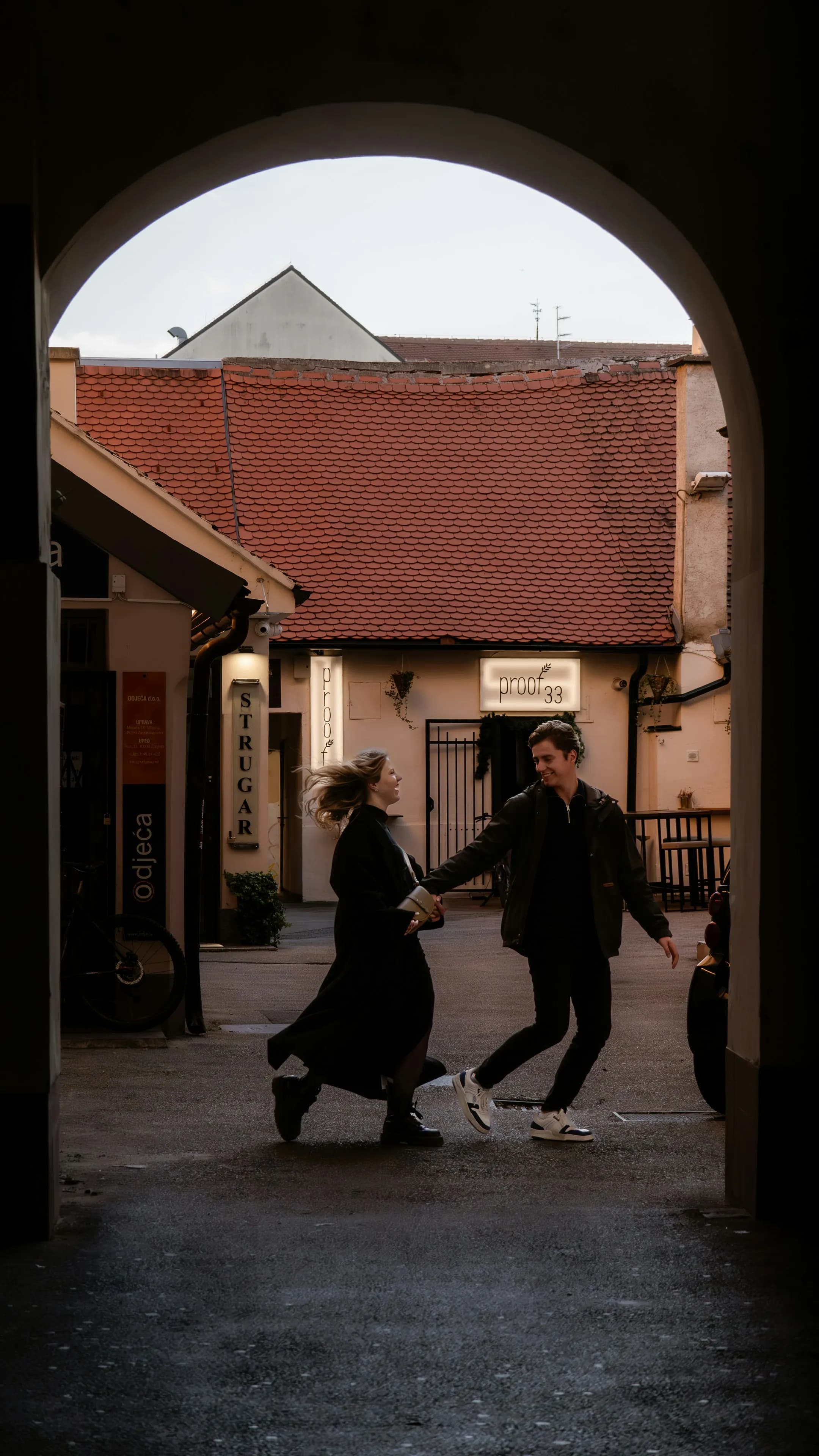 A couple holding hands runs playfully through an archway into a charming courtyard with red tiled roofs and small shops, lit by soft, natural daylight—capturing the romance found on Custom Croatia Tours.
