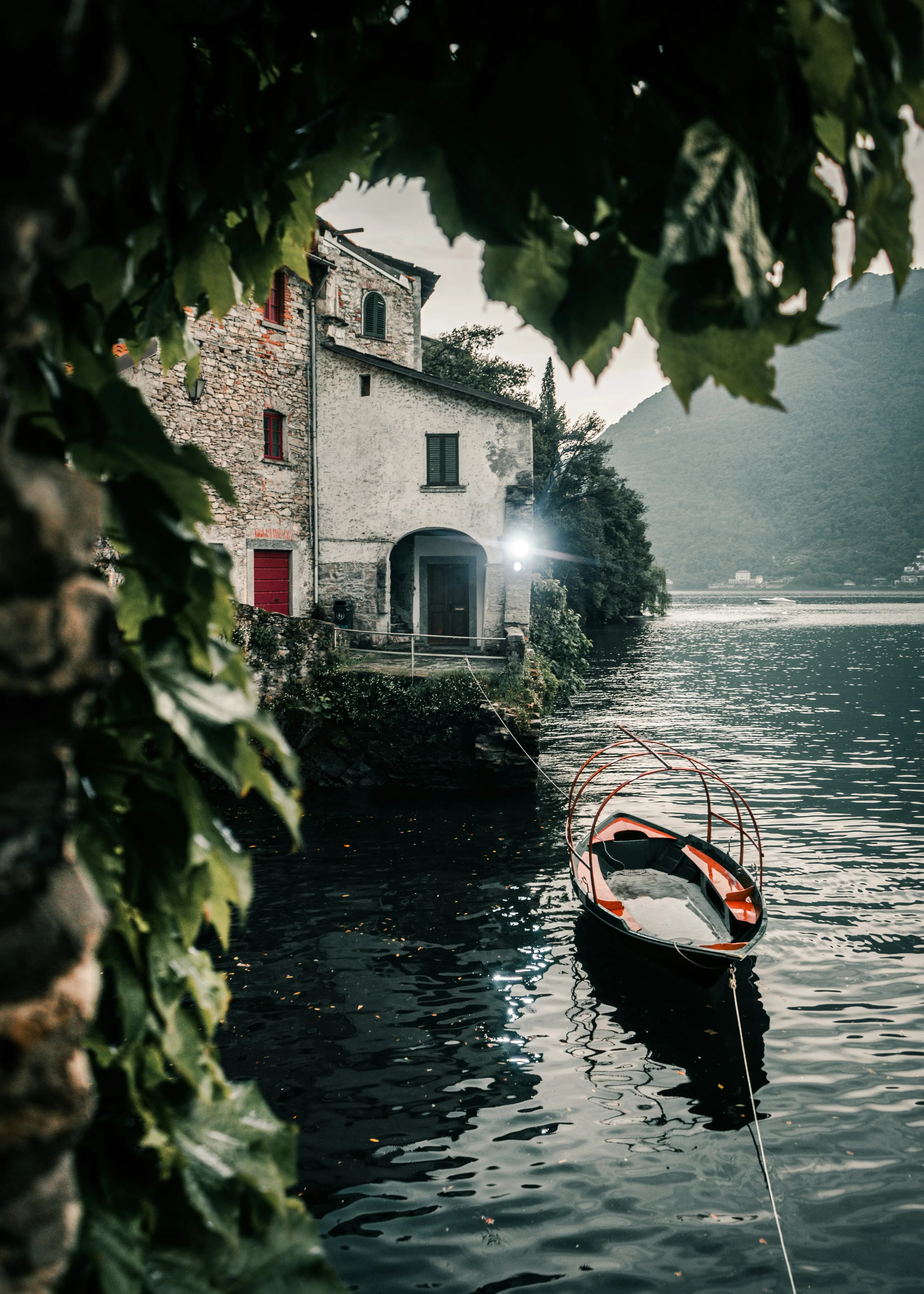 A small wooden boat is tied to the shore beside an old stone house on a calm lake, surrounded by lush greenery and misty mountains—evoking the spirit of adventure found in Vikings in Dublin 2025 Travel Packages. Leaves frame the serene scene.