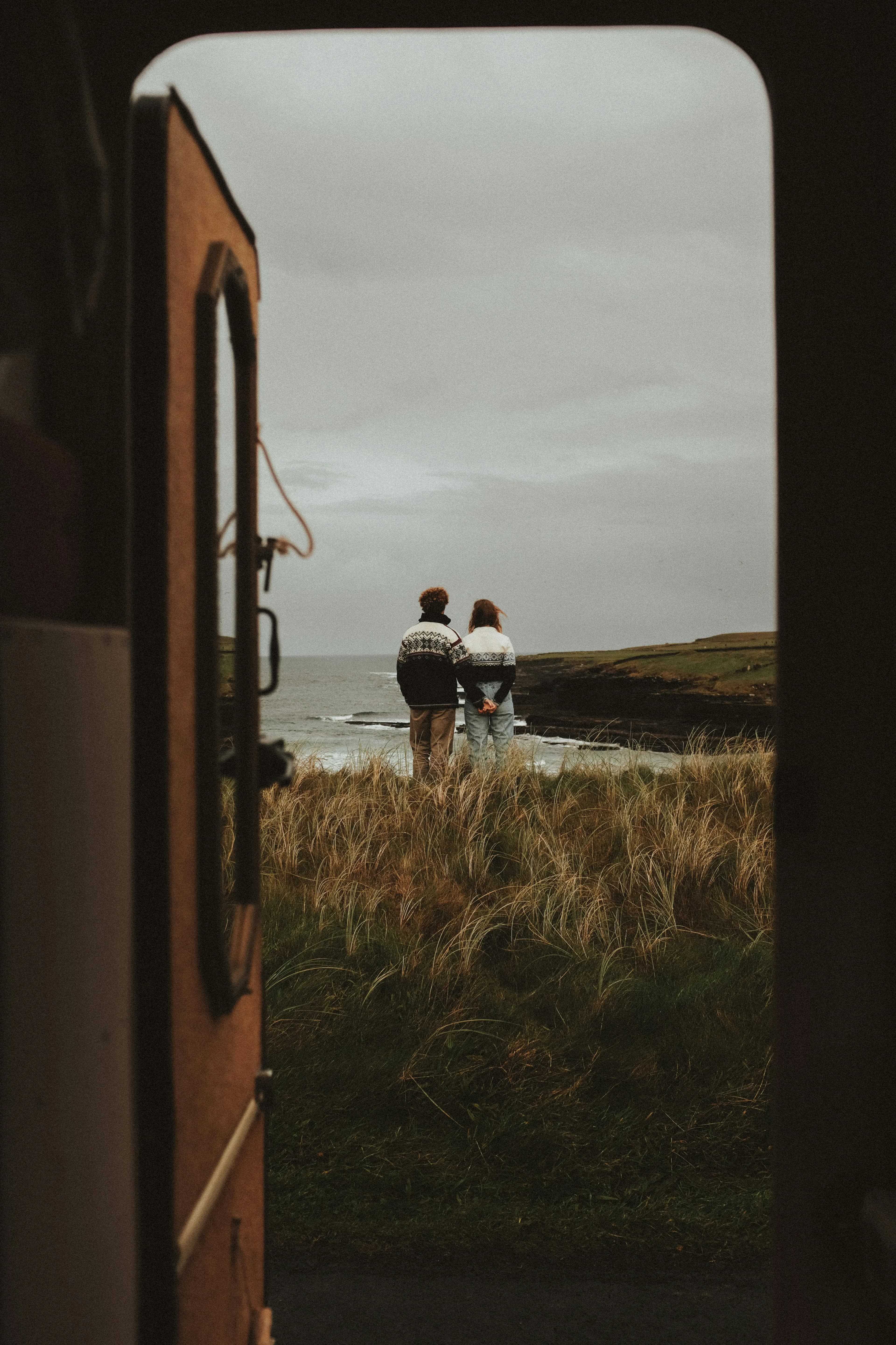 A couple stands close together on grassy terrain, facing the ocean as seen from the open door of a vehicle under a cloudy sky, capturing the romance of an Italy and Greece Honeymoon Itinerary.