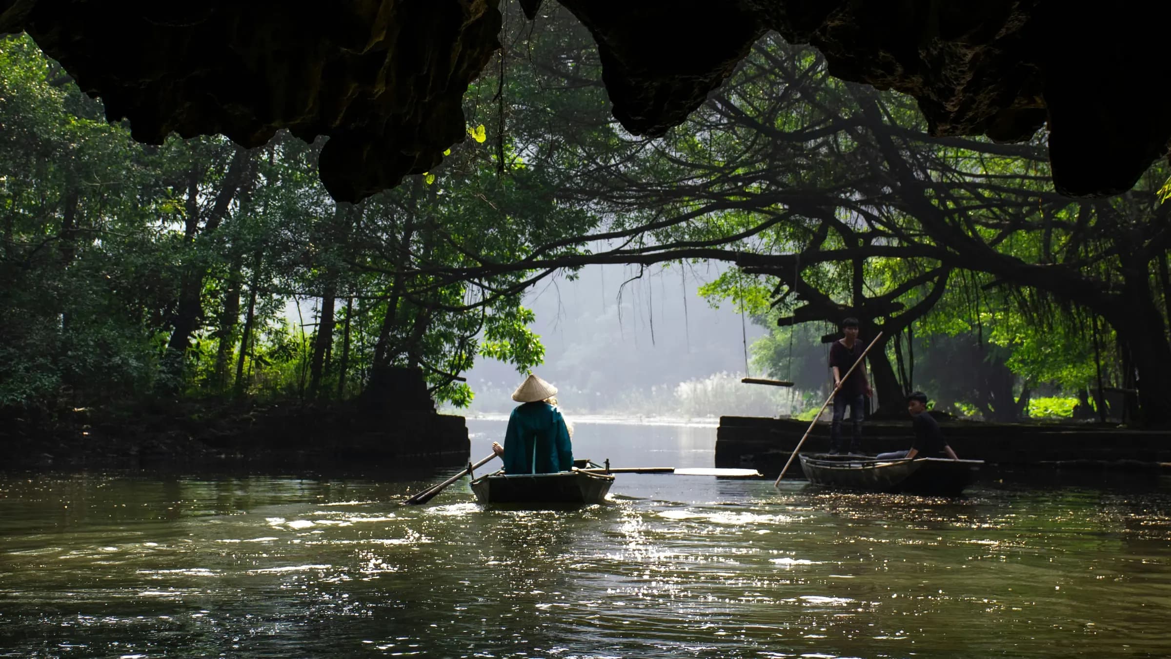 A rowing boat glides out of a limestone cave onto a green river in Trang An, northern Vietnam, framed by overhanging trees.