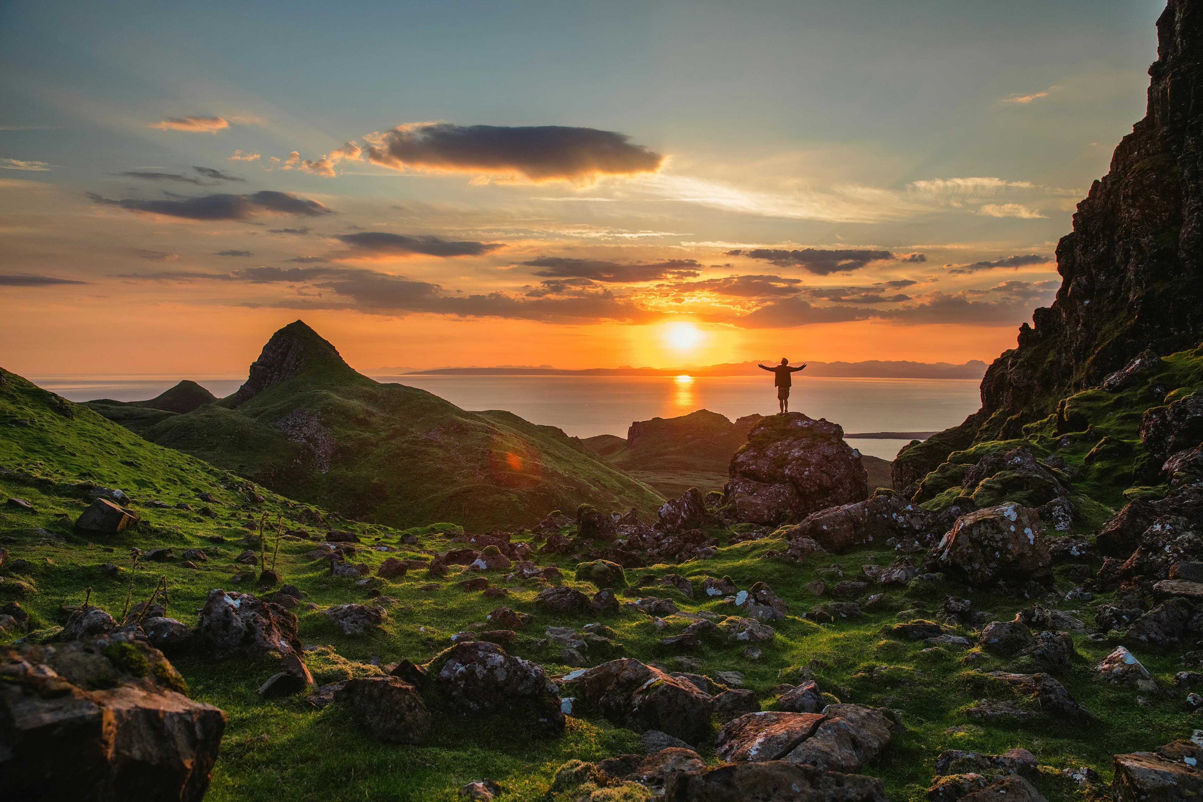 A person stands on a rocky hill with arms raised, surrounded by green hills and rocks, watching a colorful sunset over the sea and distant mountains.