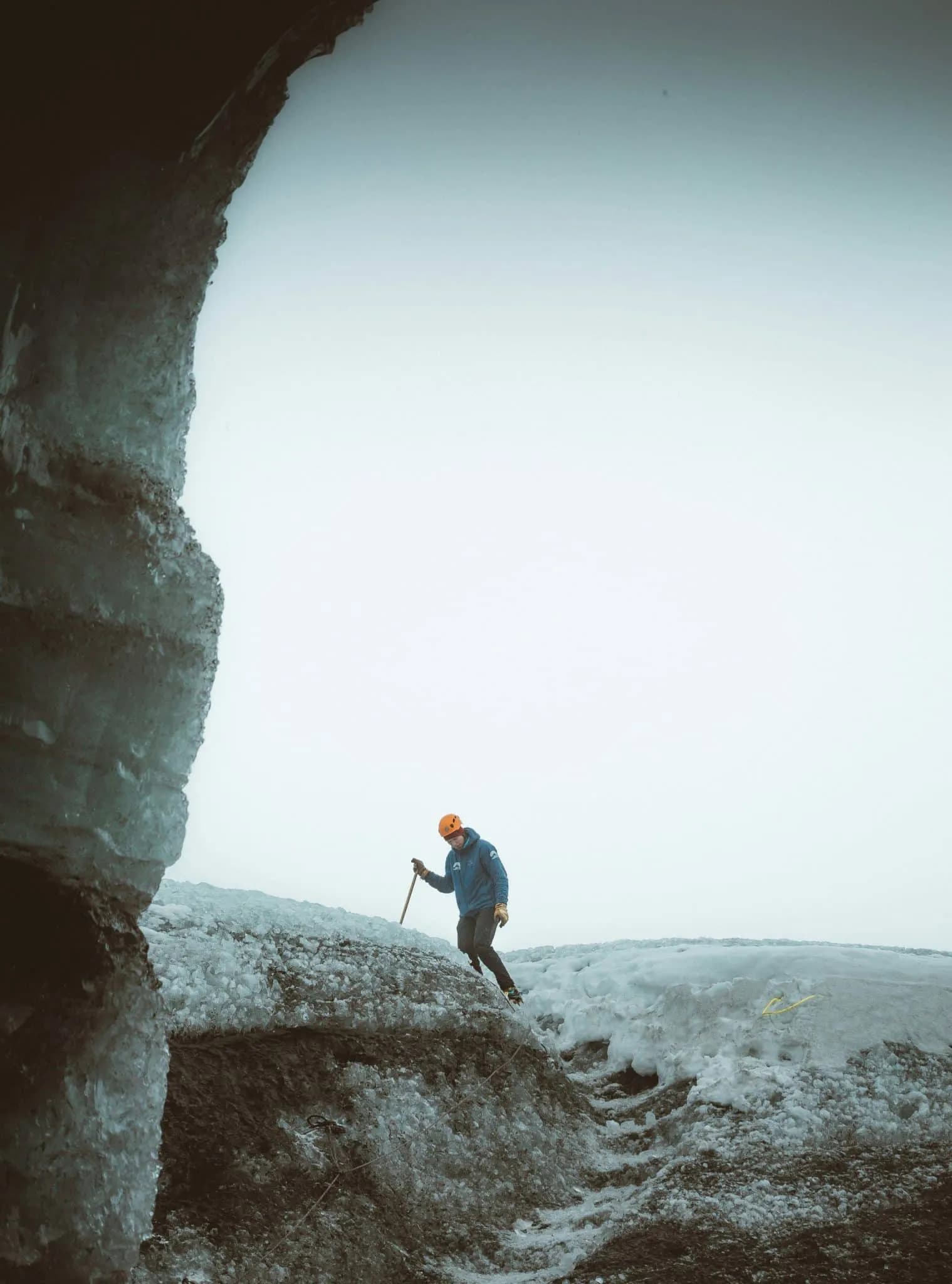 A person wearing an orange helmet and blue jacket stands on an icy terrain, holding a walking stick. The sky is overcast and a large ice formation is partially visible on the left.