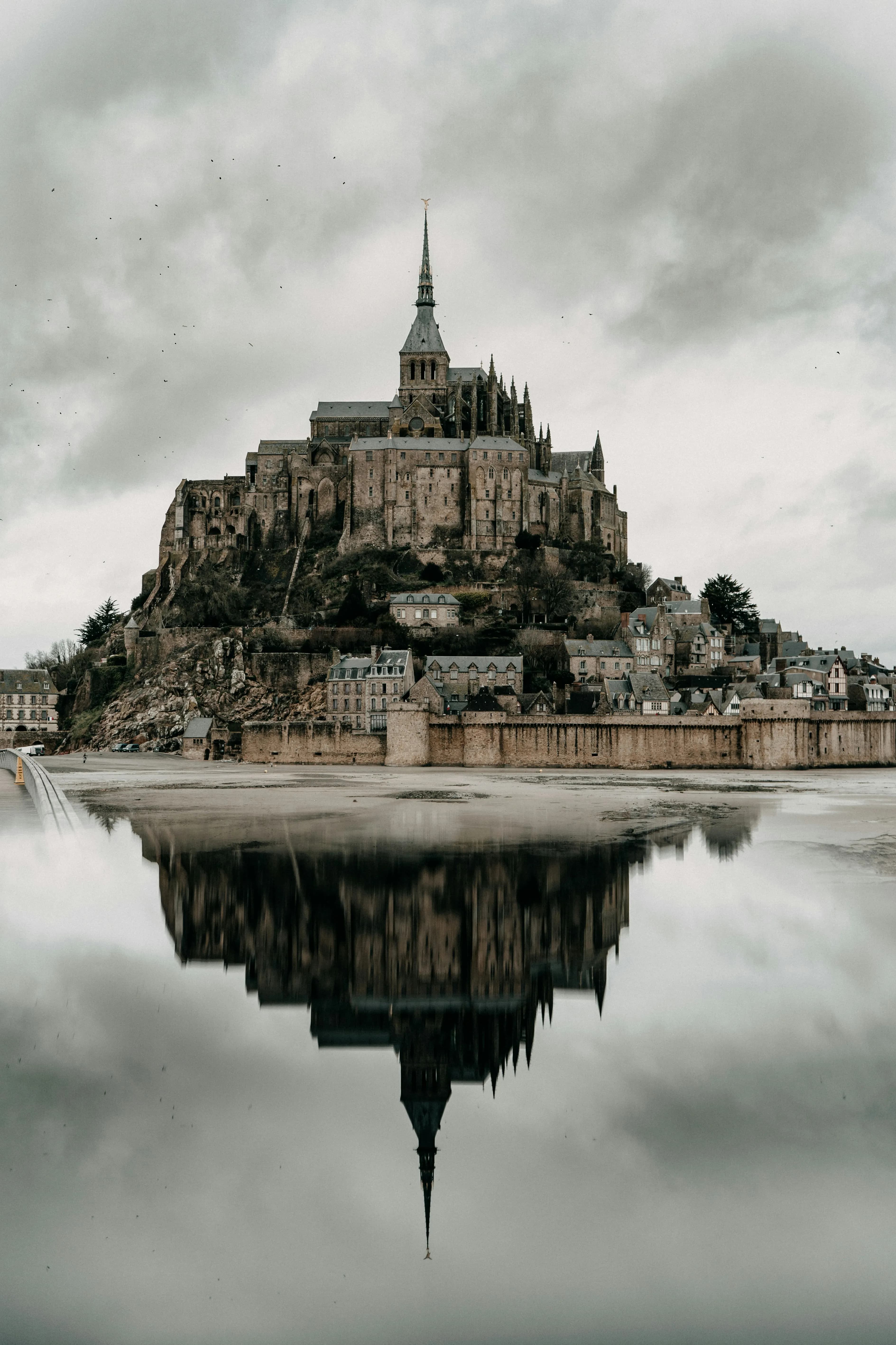 A medieval abbey sits atop a rocky hill, surrounded by small village buildings, with its reflection visible in calm water under a cloudy sky.