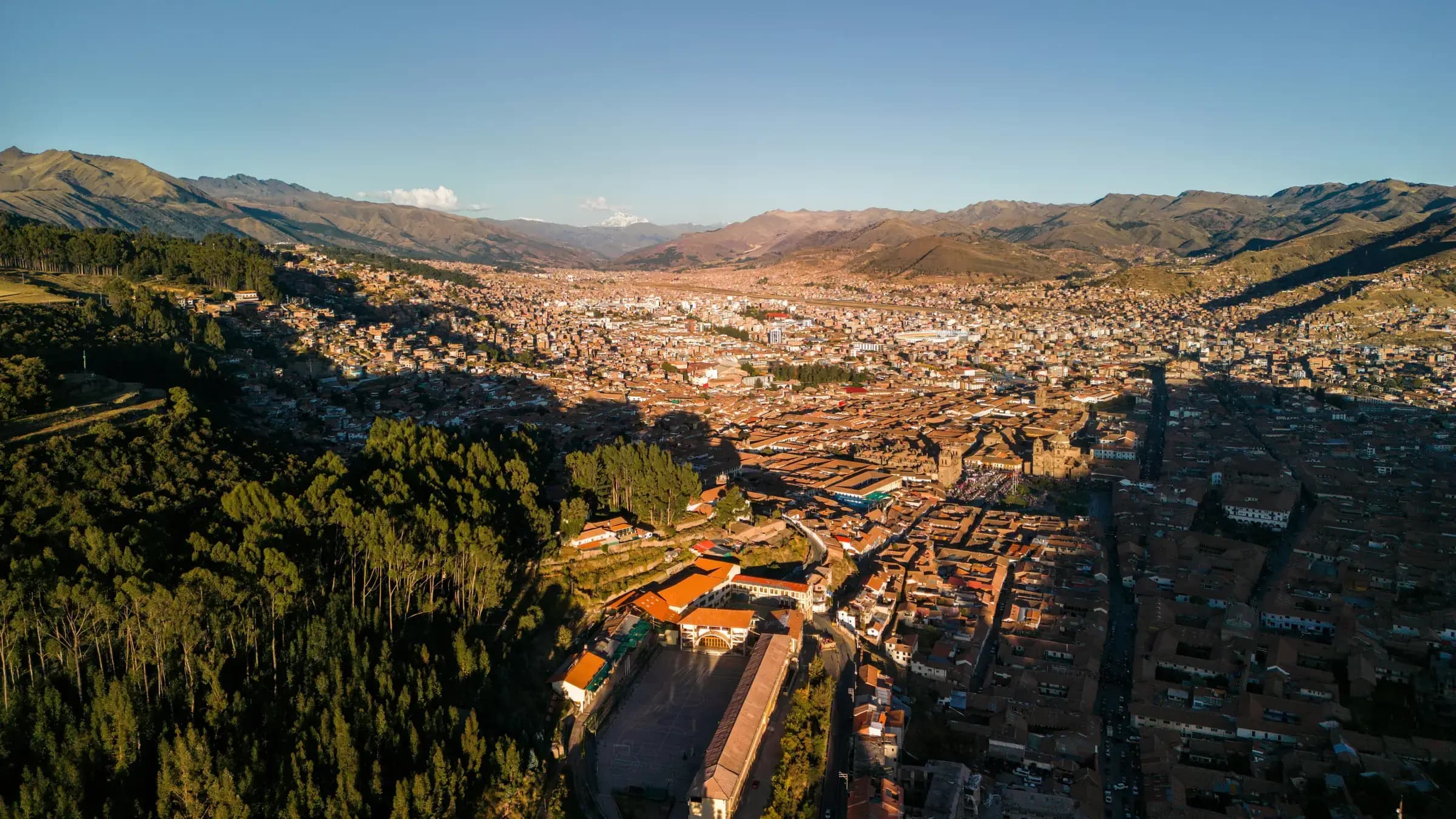 Aerial view over Cusco at golden hour, terracotta rooftops filling the valley floor with the cathedral and Plaza de Armas at centre and Andean peaks beyond.