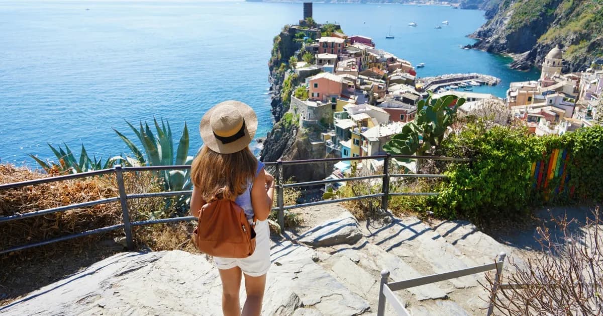A solo traveler walking down a tight, cobblestone stairway along the edge of an Italian cliff overlooking the ocean.