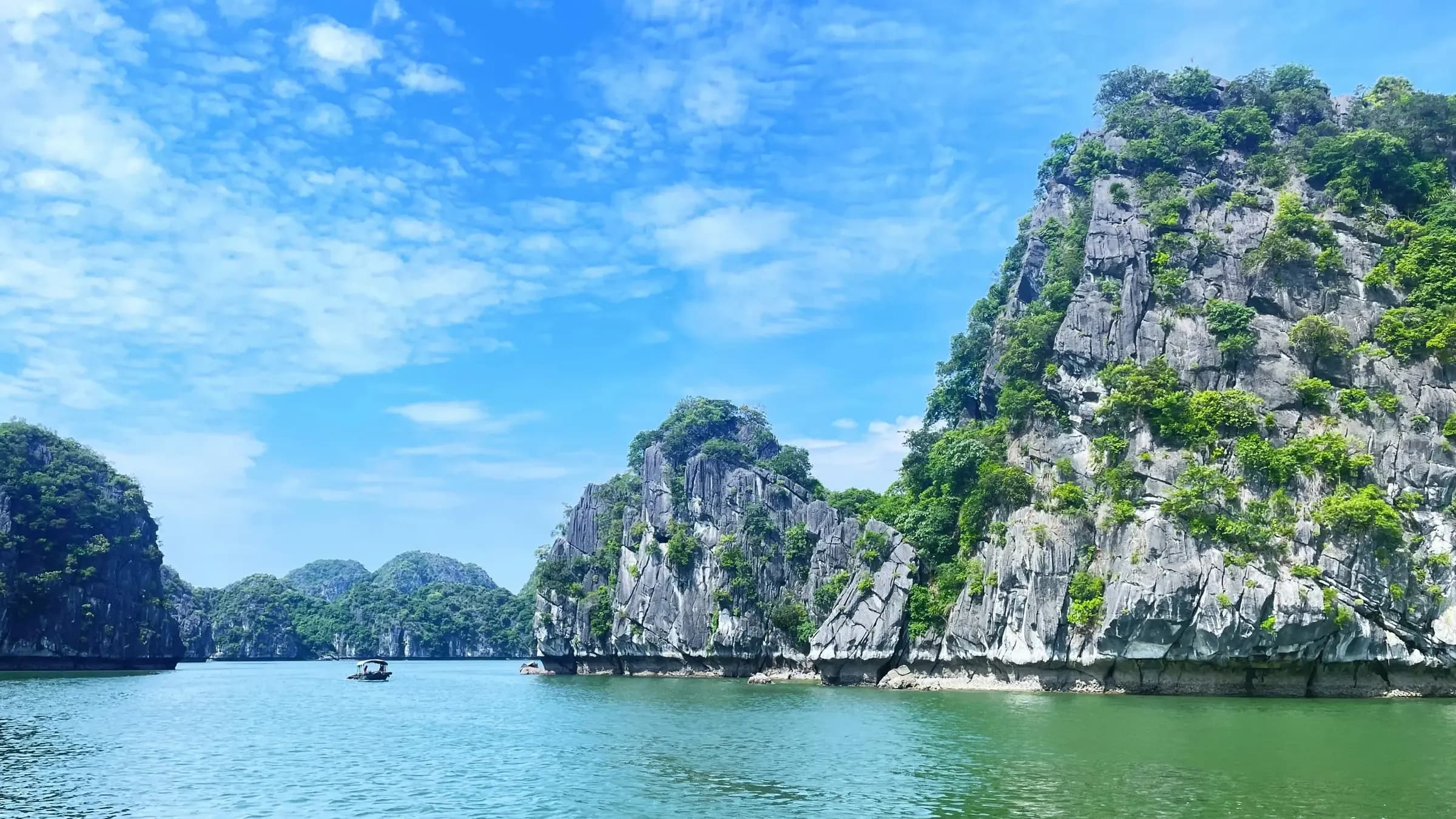Forested limestone karsts rising from calm turquoise water under a blue sky in northern Vietnam's bay country.
