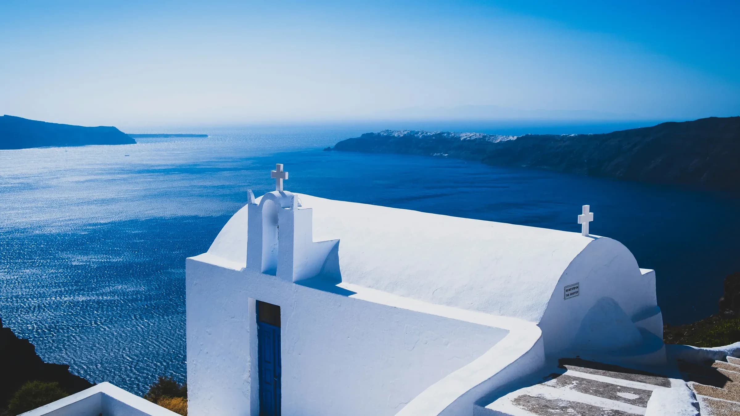 A whitewashed Cycladic chapel with a blue door overlooking the deep blue Aegean and the Santorini caldera.