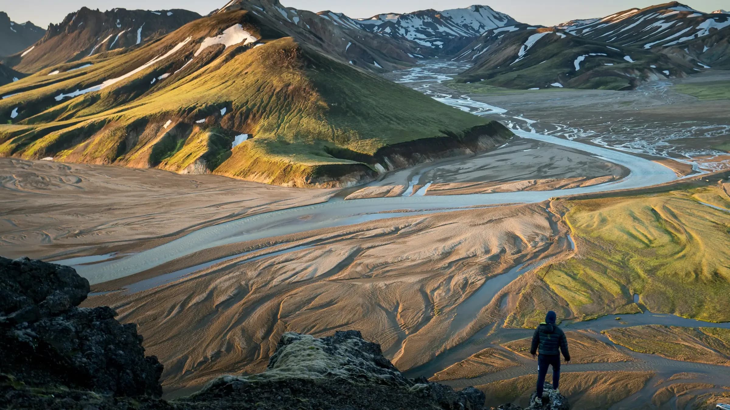 A hiker overlooking braided glacial rivers and colored rhyolite mountains in Iceland's highlands.
