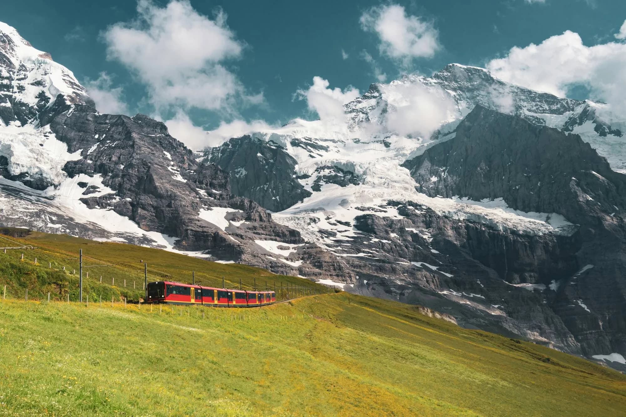 A red Jungfrau Railway train climbs a green Alpine meadow beneath snow-covered peaks and glaciers in the Bernese Oberland.