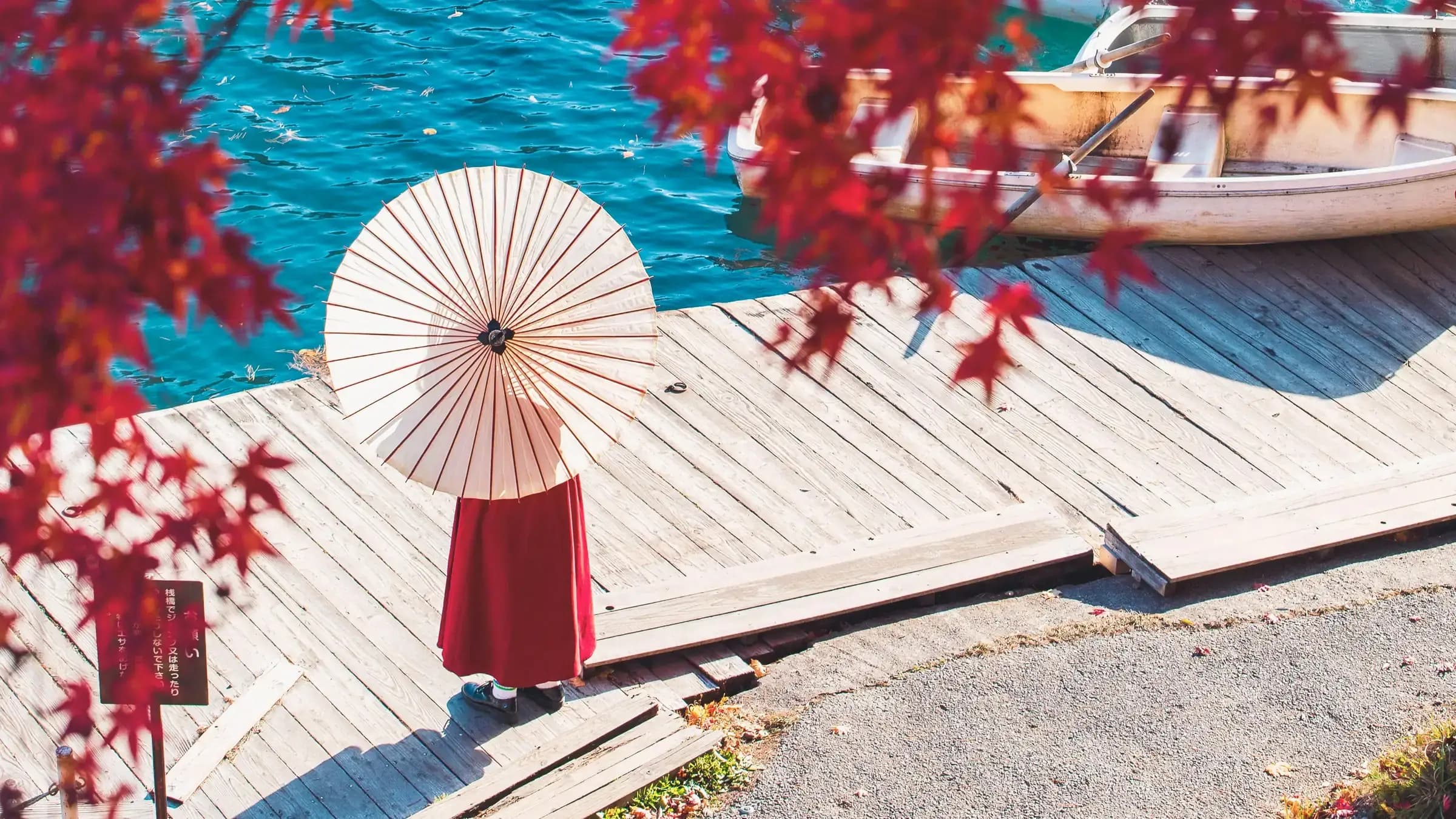 A woman in a red hakama holding a white paper parasol on a wooden jetty by bright blue water, framed by vivid red maple leaves with a rowing boat moored beyond.