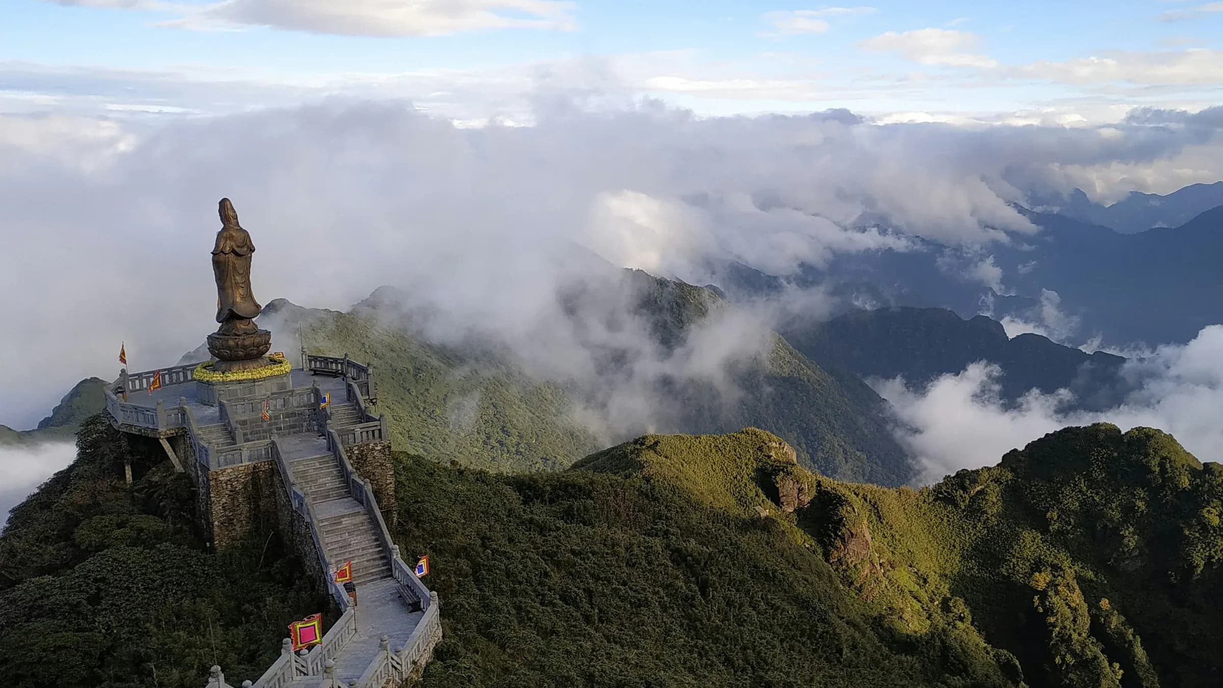 A large Buddha statue and stone stairway on a green mountain summit above the clouds in northern Vietnam.