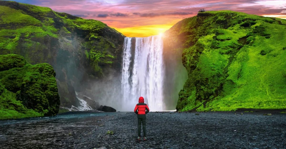 A traveler in a red jacket stands on black gravel facing Skógafoss, a wide waterfall pouring between moss-green cliffs under a sunset sky in South Iceland.