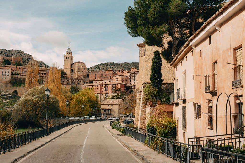 Scenic view of a winding road leading to a historic town with rustic buildings and a prominent church tower. Trees with autumn foliage line the road, and the background features hills under a partly cloudy sky. Albarracín, Spain