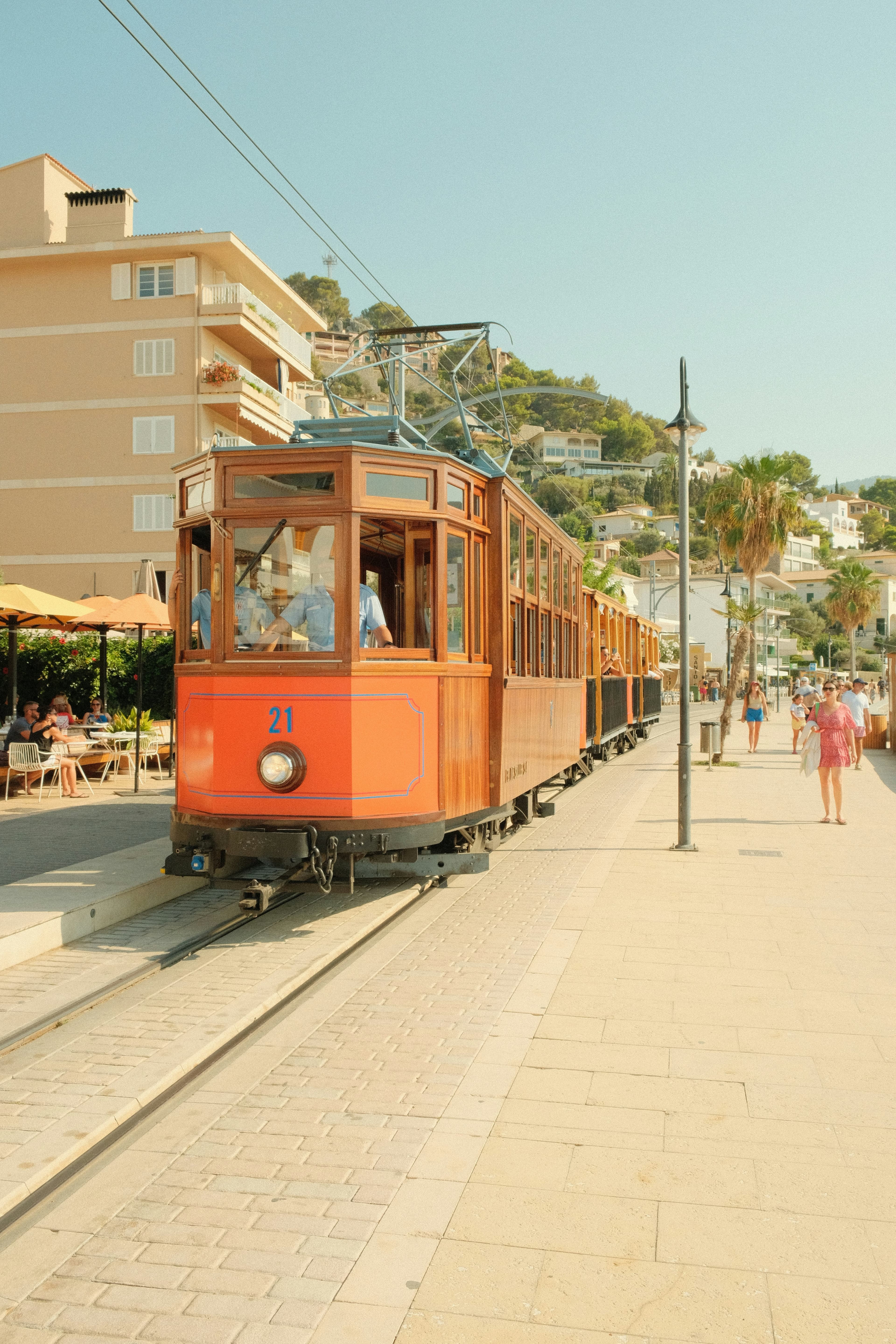 A vintage wooden tram moves along tracks beside a sunny promenade, with people walking and sitting at outdoor café tables—reminiscent of relaxing after visiting castles in Scotland to tour for free. Mediterranean style buildings and palm trees complete the scene.