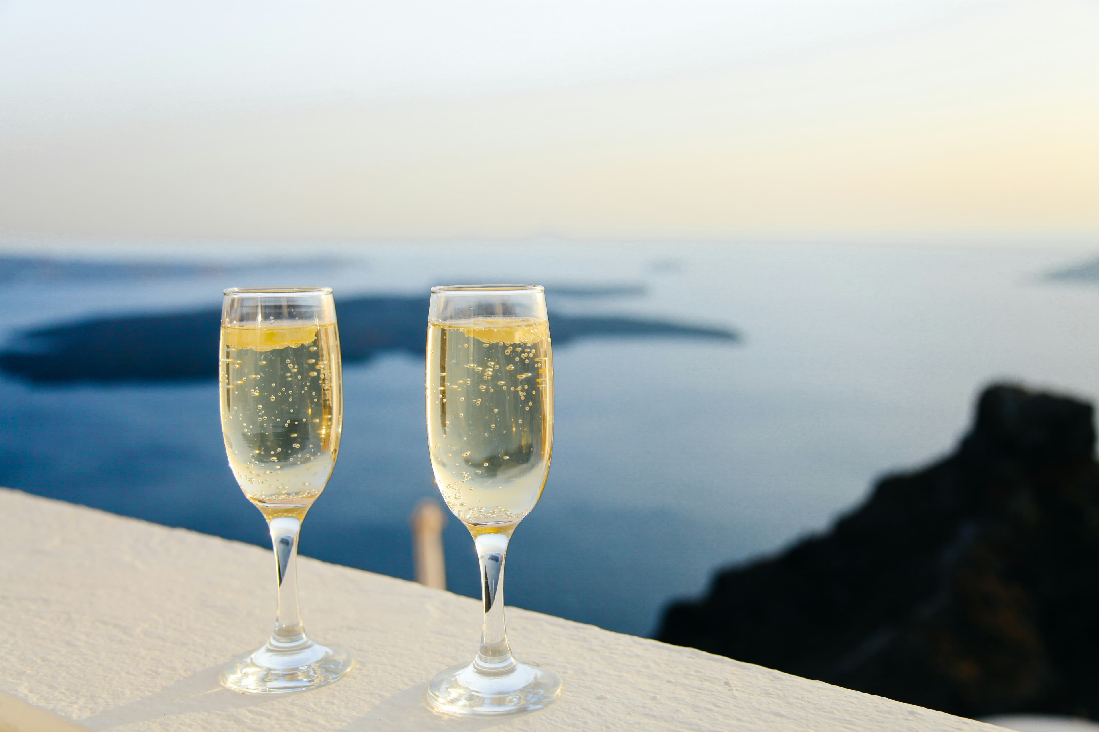 Two glasses of sparkling wine sit on a white ledge, with a scenic, blurred view of the sea and distant islands at sunset—capturing the romance found in unforgettable Spain and Portugal Honeymoon Packages.
