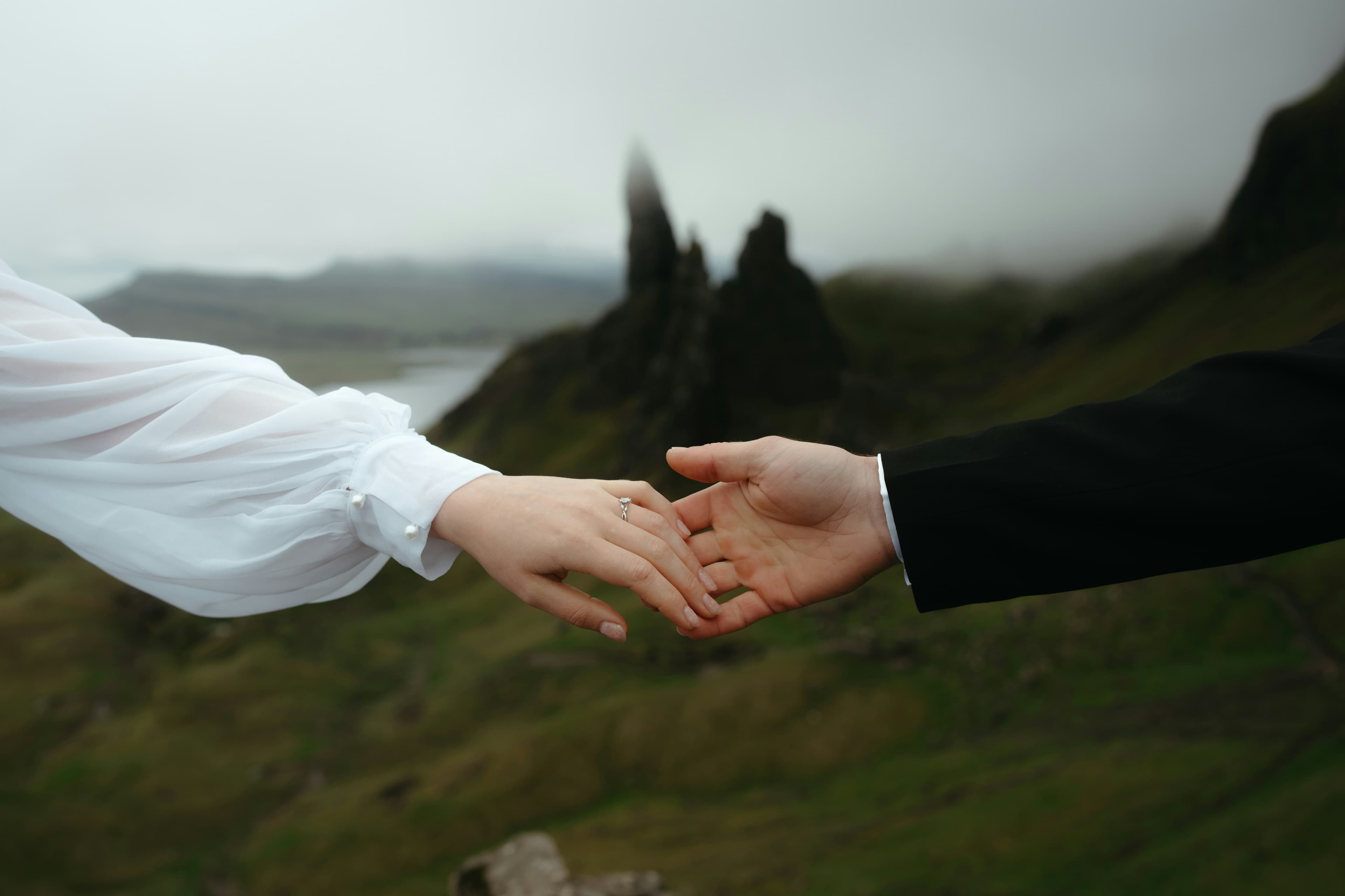 Two people reach out to hold hands, one in a white sleeve and the other in black, framed by a misty, mountainous landscape—capturing the romance of Custom Scotland Tours.