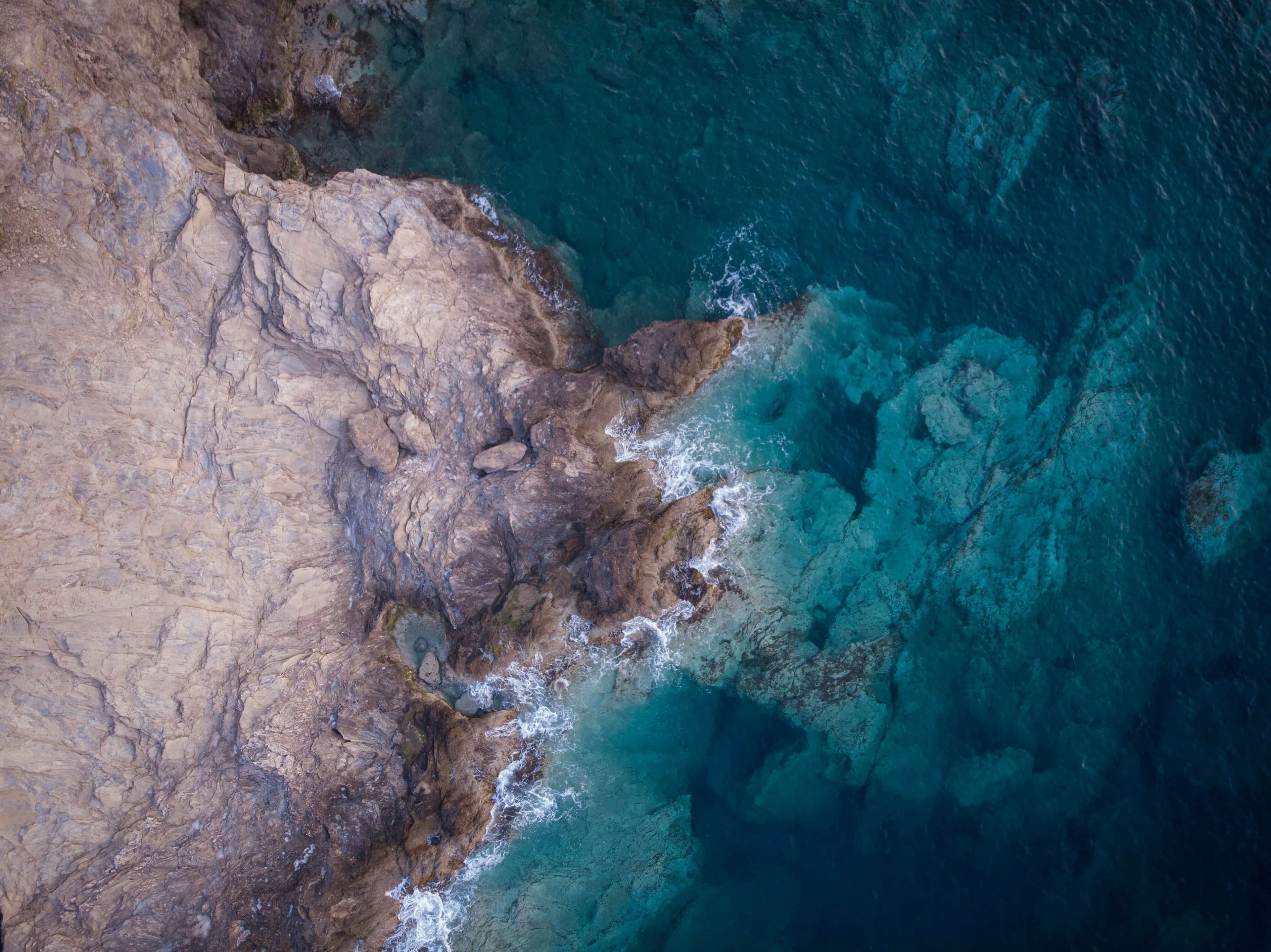 Aerial view of a rocky coastline with waves crashing against the shore and clear blue green ocean water, reminiscent of the intensity seen in the Notre Dame Navy Rivalry, revealing striking underwater formations near the coast.