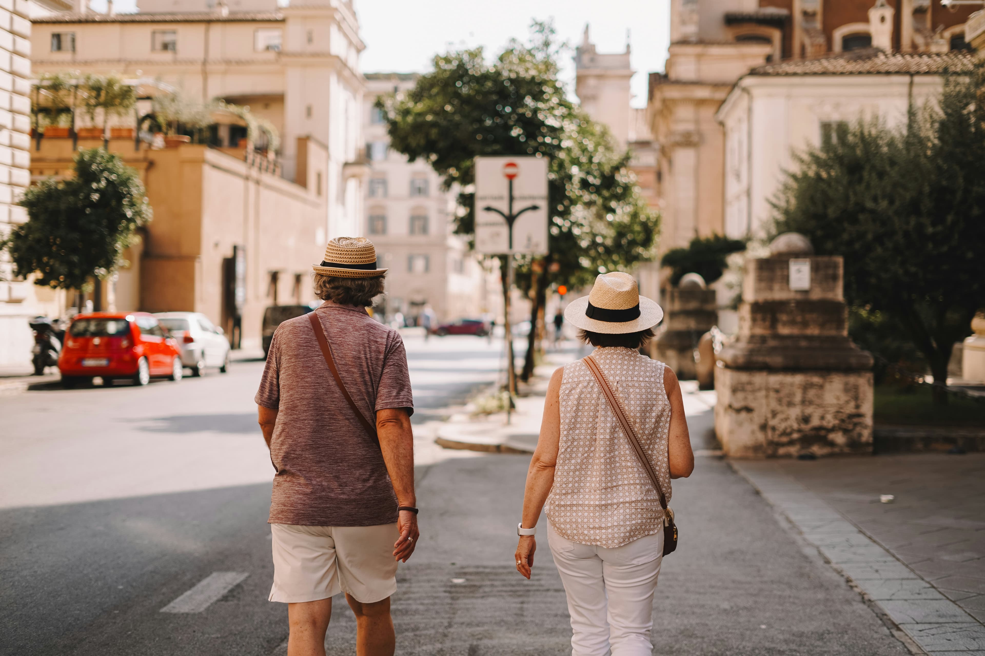 An elderly couple wearing summer hats walks together down a sunny, quiet street lined with historic buildings and trees, reminiscing about their dream trip to a Glass Igloo in Iceland as they enjoy their leisurely stroll.