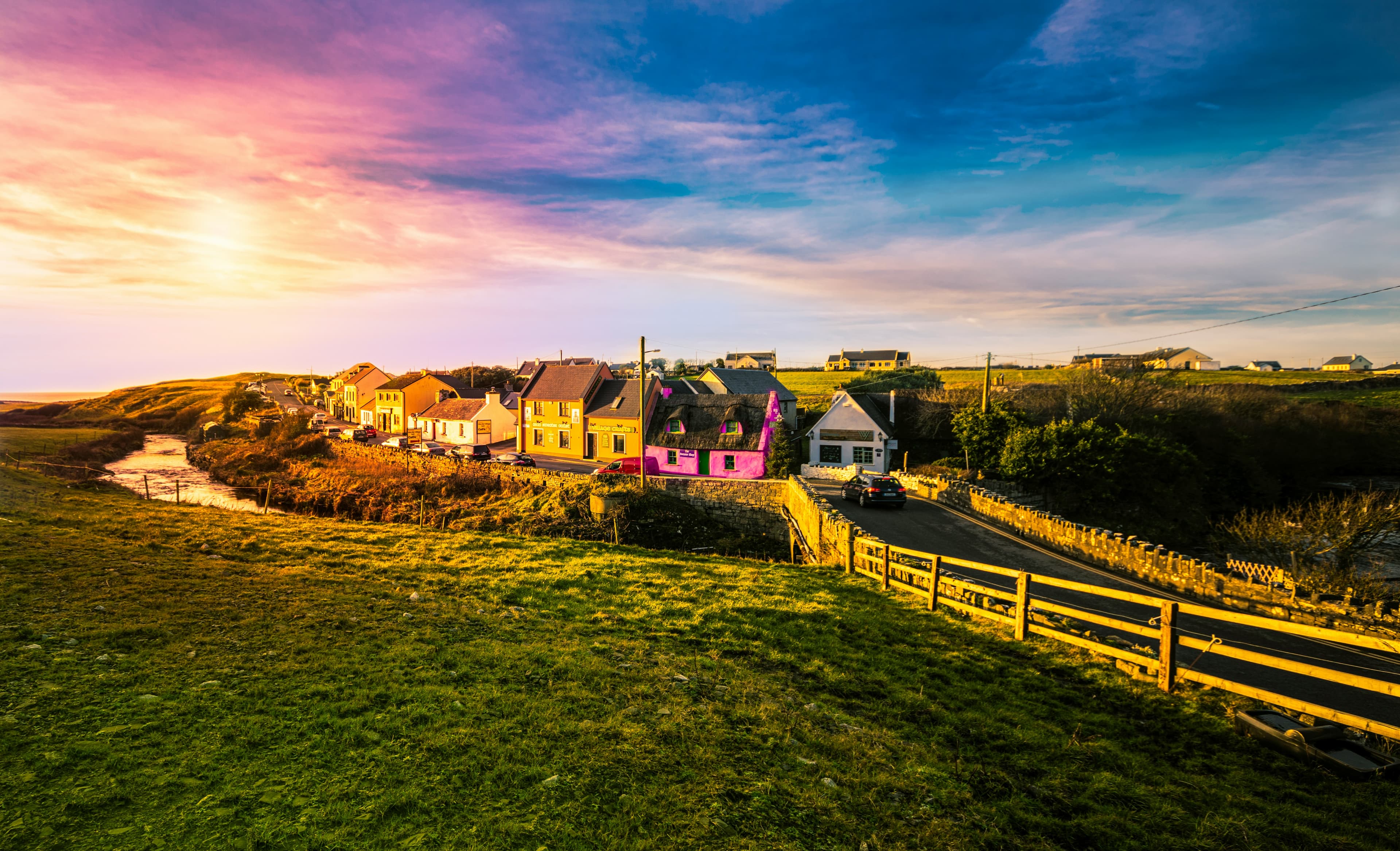 A row of colorful houses, including a bright pink one, lines a small road in a rural Irish village at sunset. A wooden fence and grassy fields surround the village, with a river running alongside—perfect for an idyllic Ireland honeymoon.