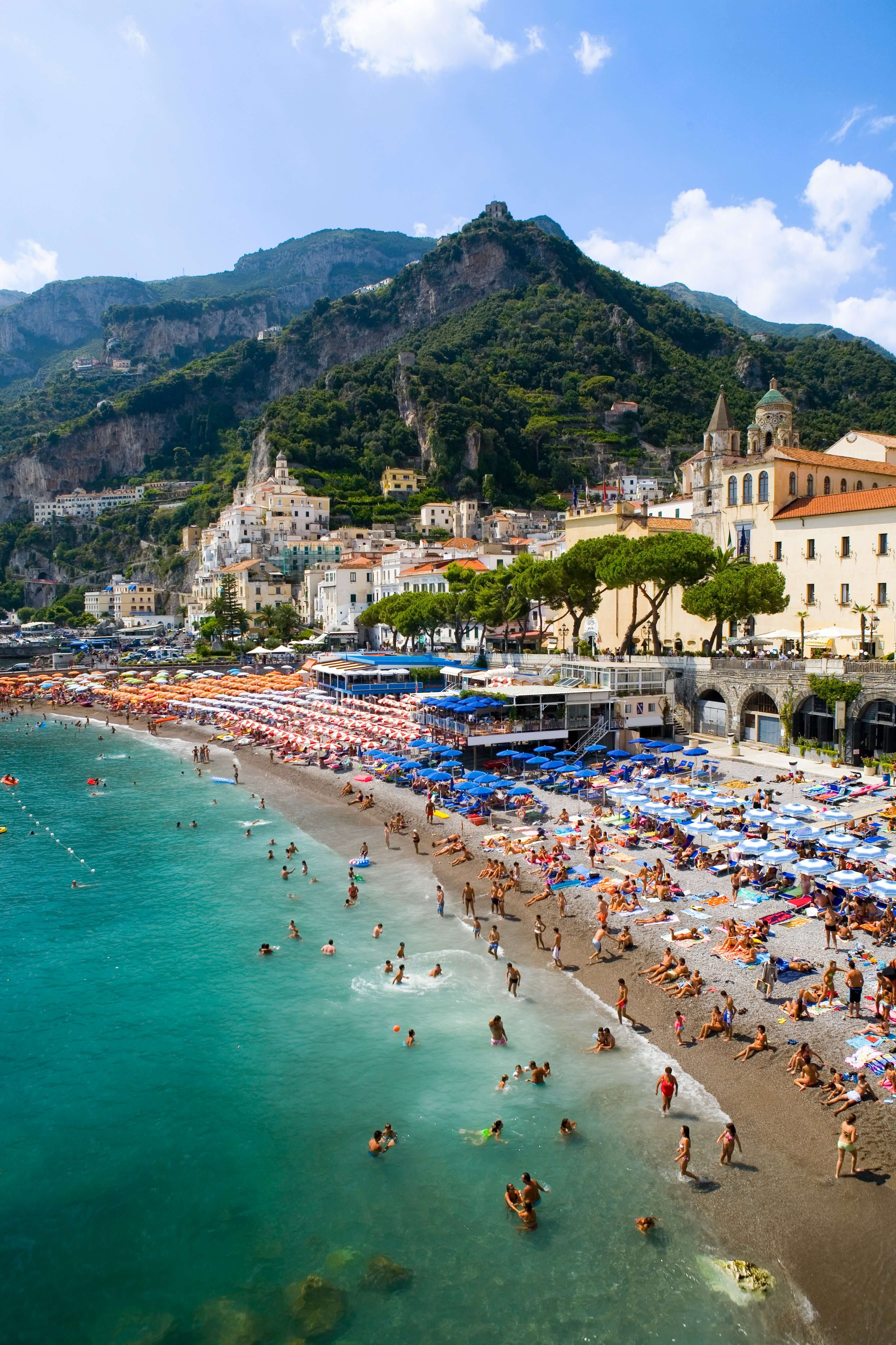 A crowded beach with turquoise water, colorful umbrellas, and sunbathers; like a stop on your Ireland Honeymoon Itinerary 10 Days, a coastal town with historic buildings and lush green mountains rises in the background under a sunny sky.